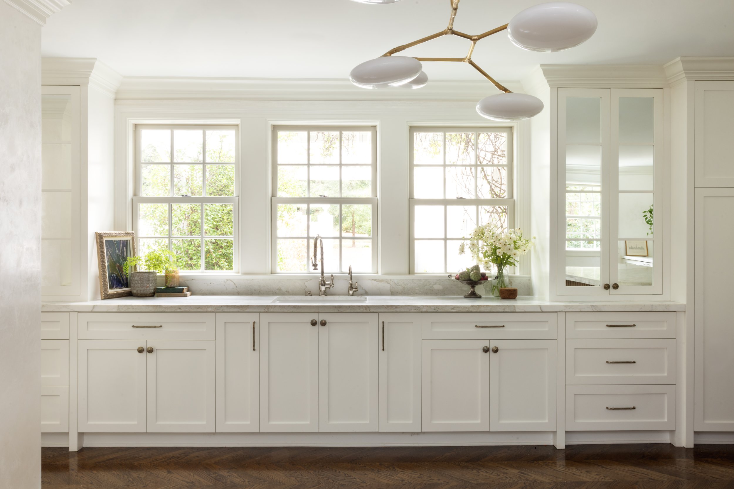 Bright kitchen with white cabinets, marble countertops, and large windows with greenery outside. Decor includes framed artwork, potted plants, and flower arrangements.