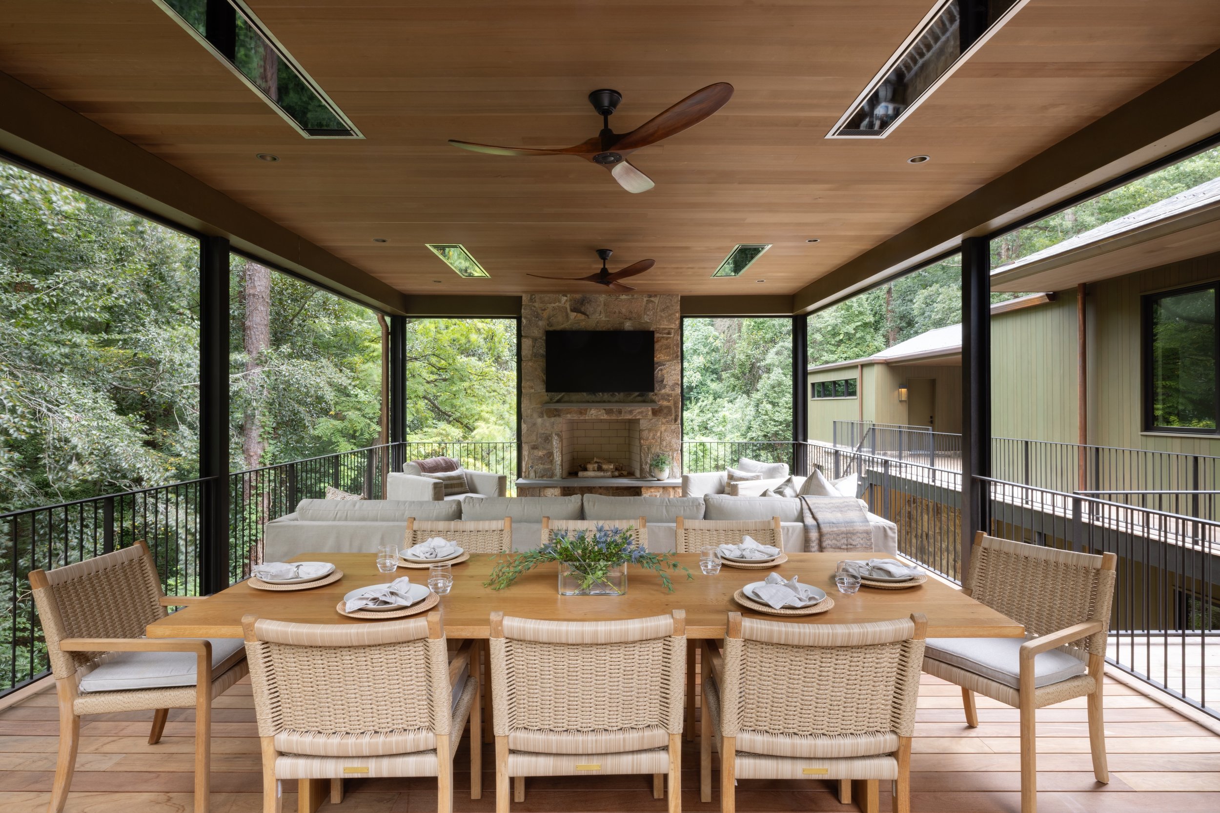Indoor dining and living area with a wooden table, nine chairs, a stone fireplace, a flat-screen TV, and a seating area, surrounded by large windows with a view of trees outside.