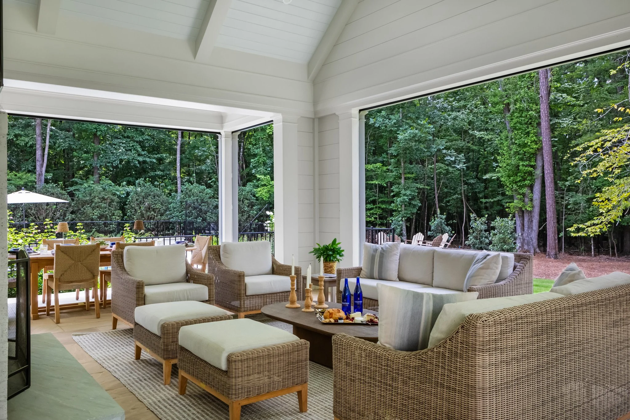 Luxury covered outdoor living room built by Will Johnson Building Company featuring comfortable seating, woven outdoor furniture, neutral cushions, overhead ceiling detail, and open views into a wooded landscape designed for relaxed entertaining.