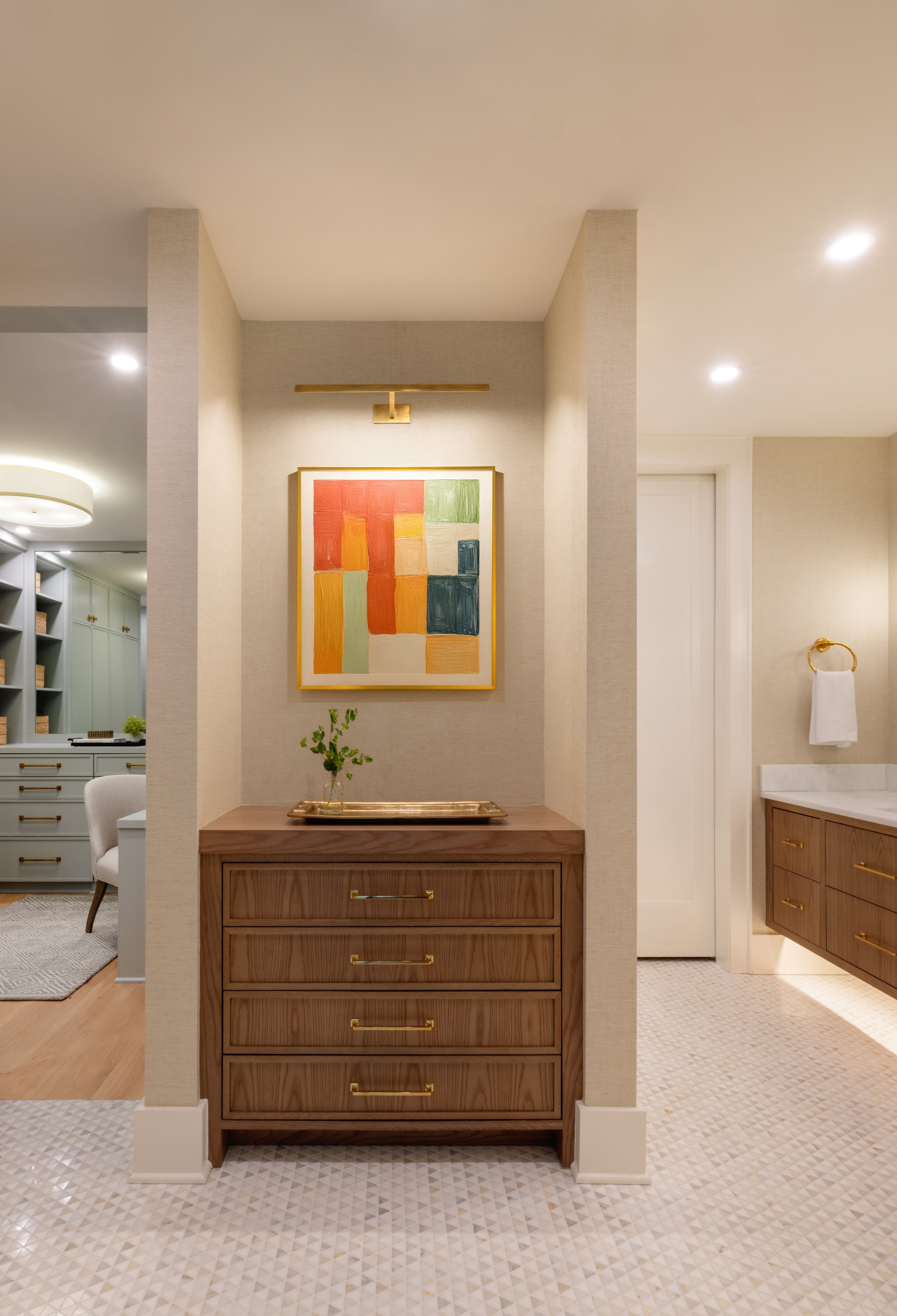 A wooden dresser with four drawers and gold handles, topped by a gold tray with a small green plant, in a well-lit room. An abstract colorful painting hangs above, with beige walls and a partial bathroom visible on the right.