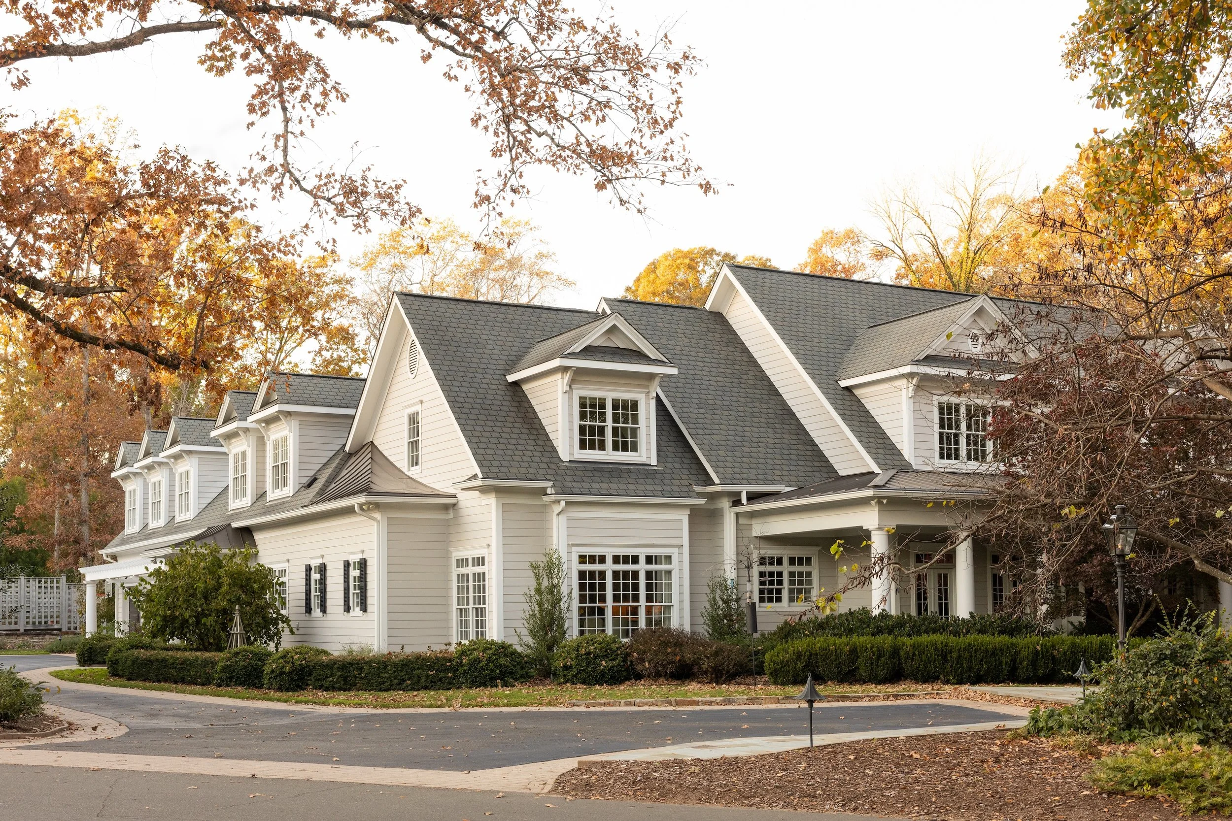 White two-story house with dormer windows, surrounded by trees with fall foliage, and a curved driveway in front.