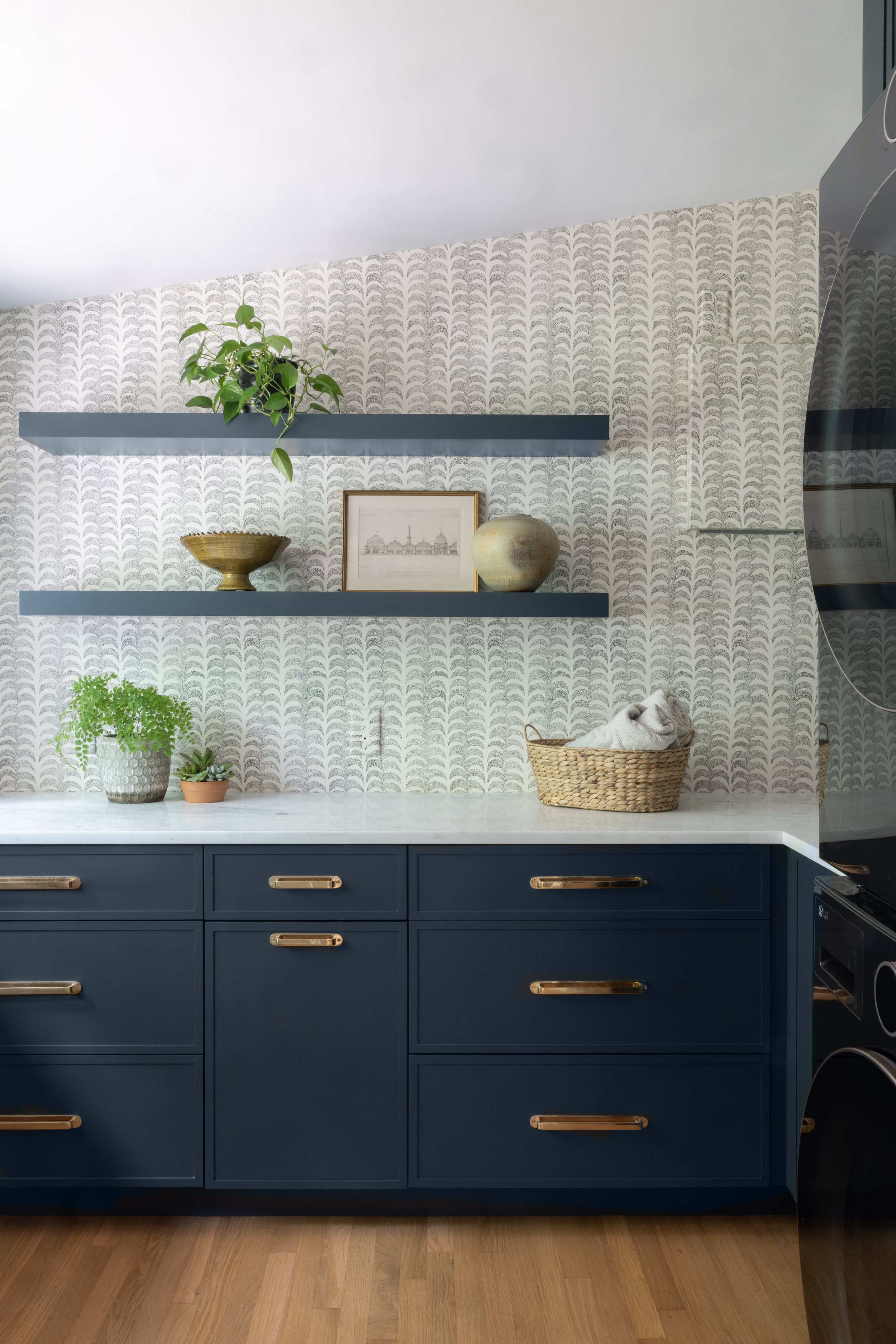 A kitchen corner with navy blue cabinets featuring gold handles, a white marble countertop, and two floating navy blue shelves decorated with potted plants, a framed picture, a bowl, and a decorative ceramic piece. The wall behind has light patterned