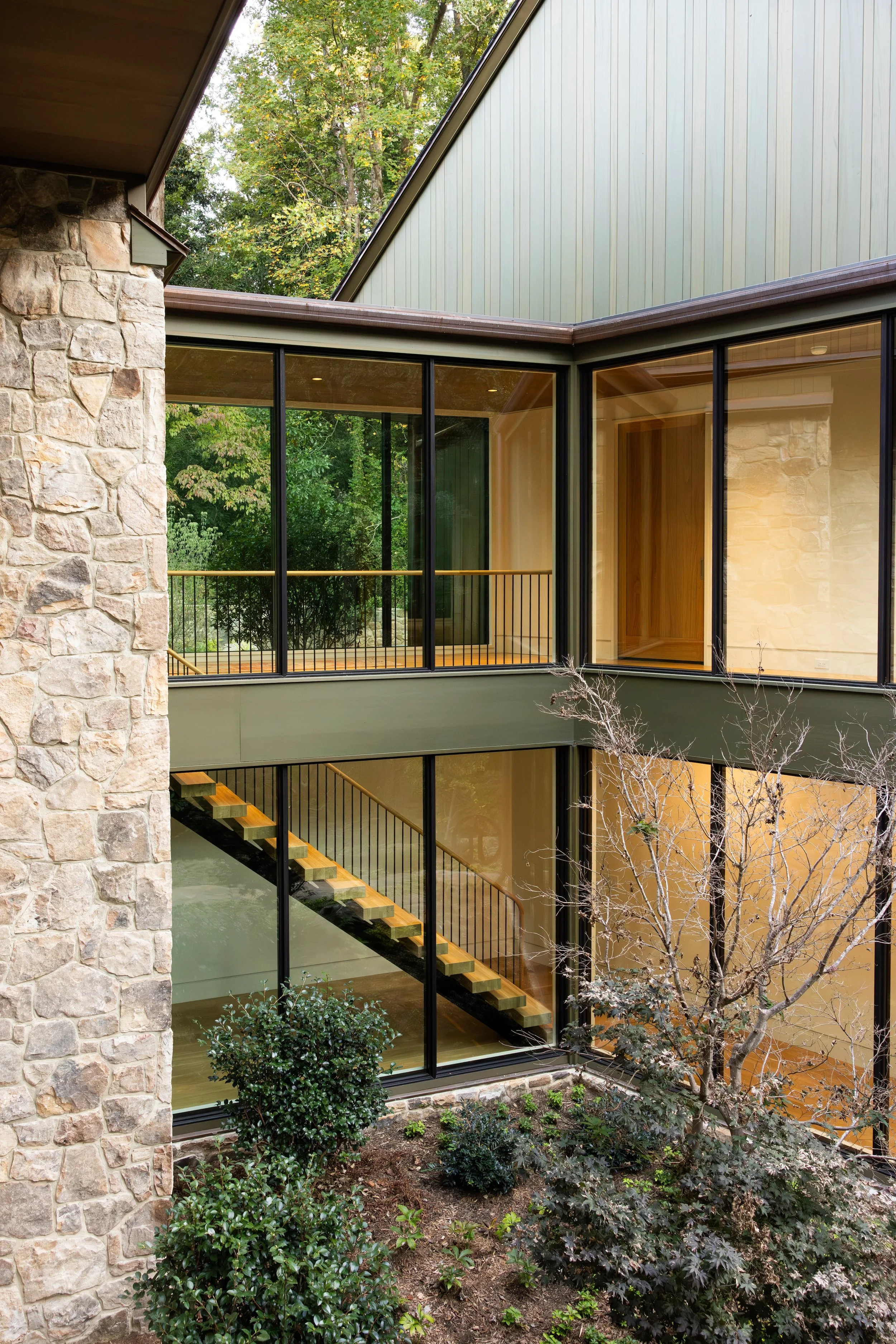 A modern multi-story building with large glass windows, a stone wall on the left, wooden staircase inside visible through the glass, and a garden with bushes and a leafless tree outside.