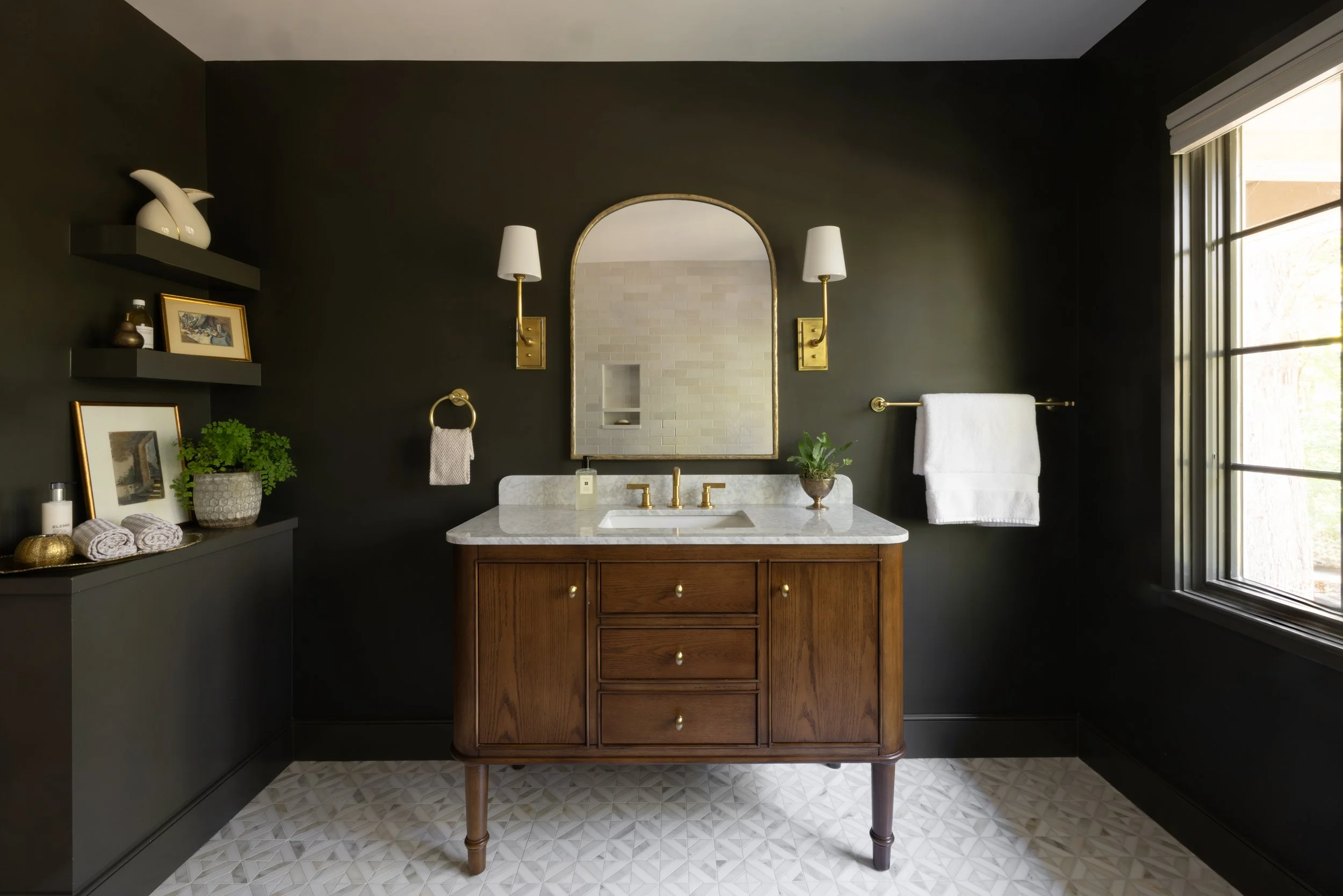Bathroom with dark walls, wooden vanity with marble top, gold fixtures, large mirror, wall-mounted lights, towel rack, window with natural light, decorative items and plants.