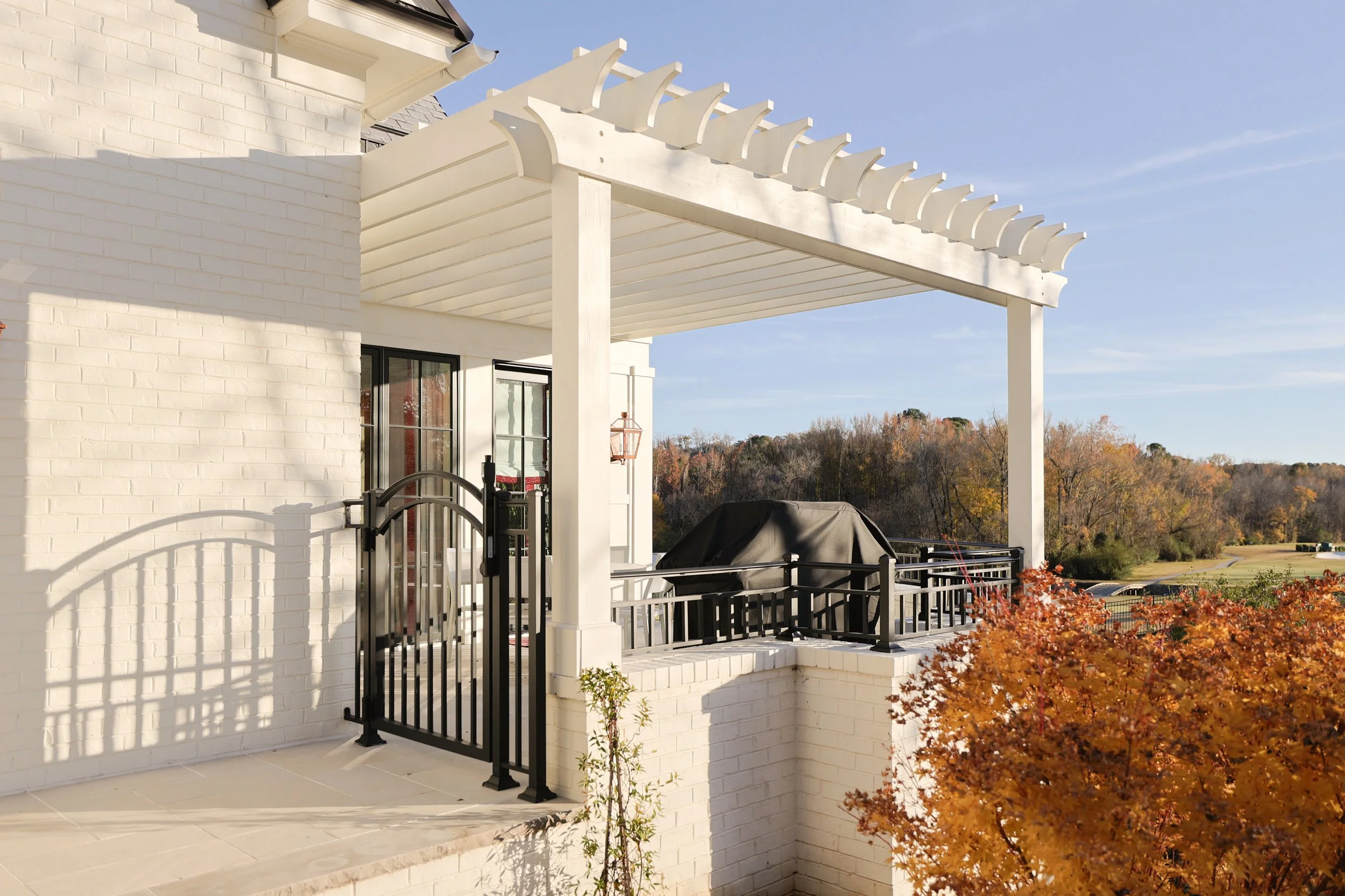 A white brick house with a black wrought iron gate and a covered porch, with a grill covered by a black cover on the patio, and autumn trees in the background.