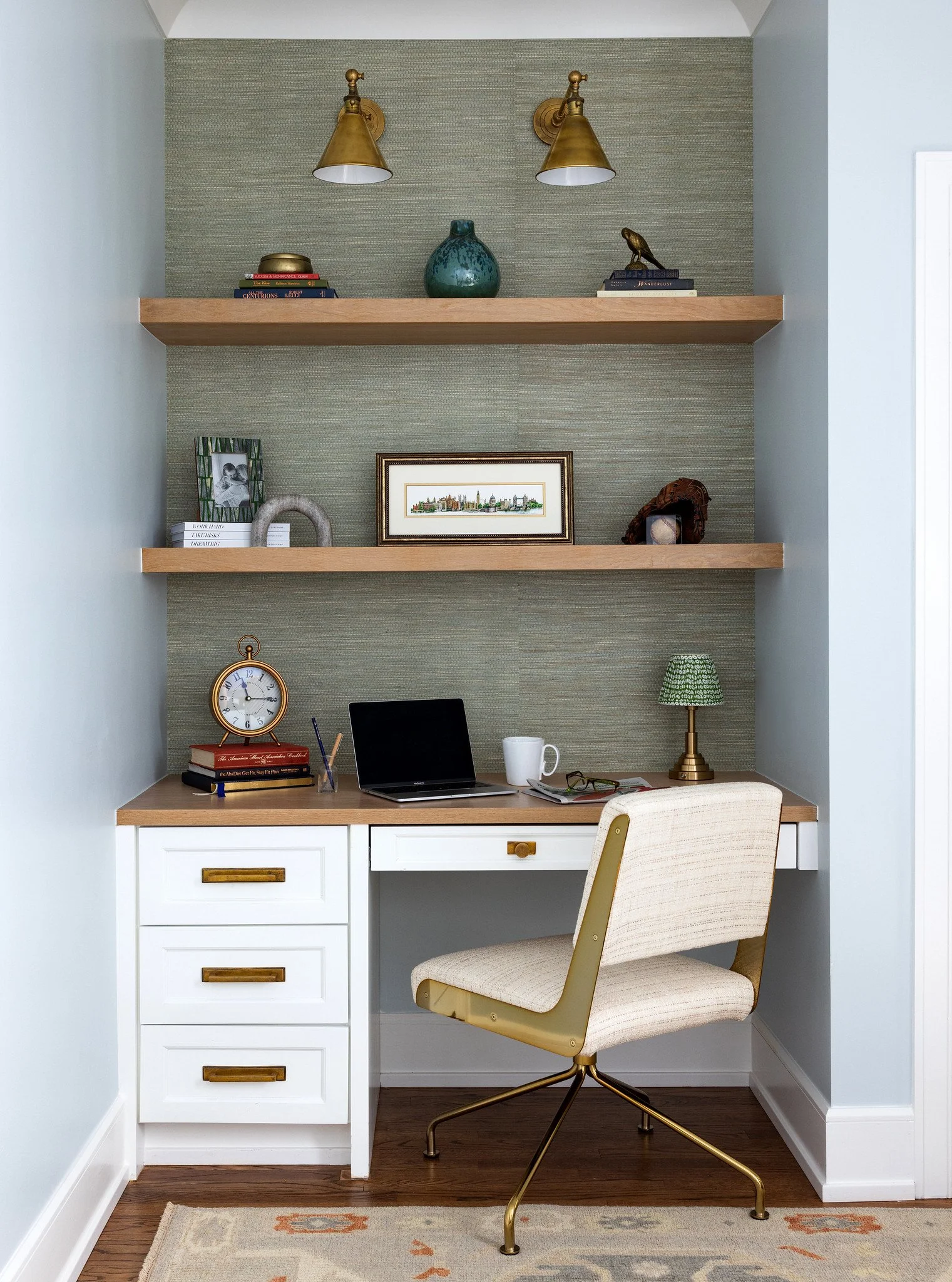 A home office with a white desk, beige upholstered swivel chair, two wall-mounted gold lamps, three wooden shelves with decorative items, a laptop, books, a clock, a coffee mug, and a green table lamp.