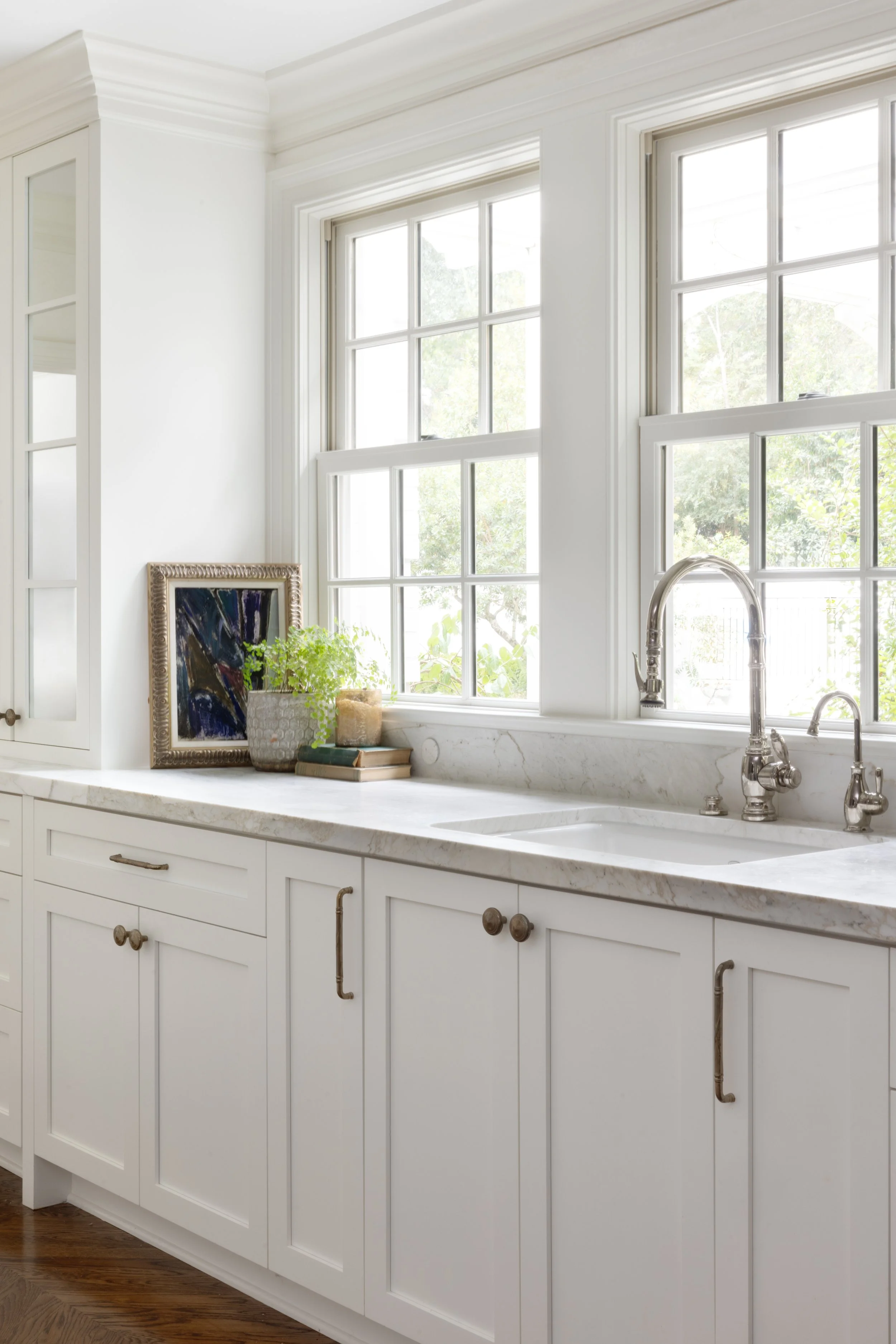 Bright kitchen window with white cabinets and marble countertop, decorated with potted plants, books, and an abstract framed artwork.