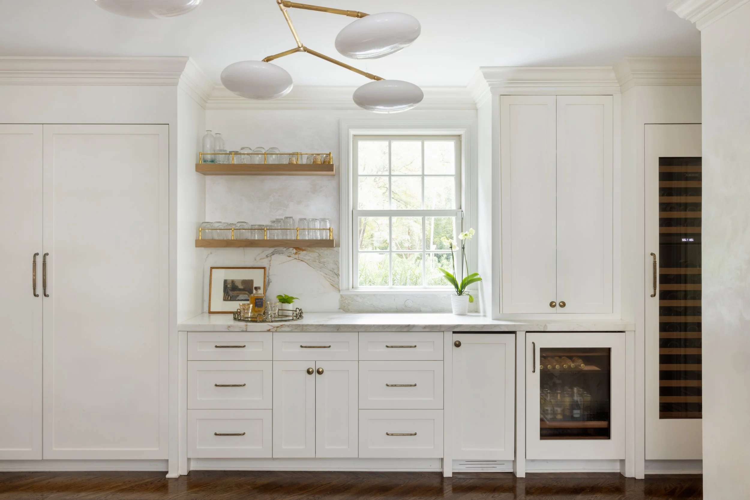 Bright kitchen with white cabinetry, marble countertops, open wooden shelves with glassware, a window with a potted orchid, and a built-in wine fridge.