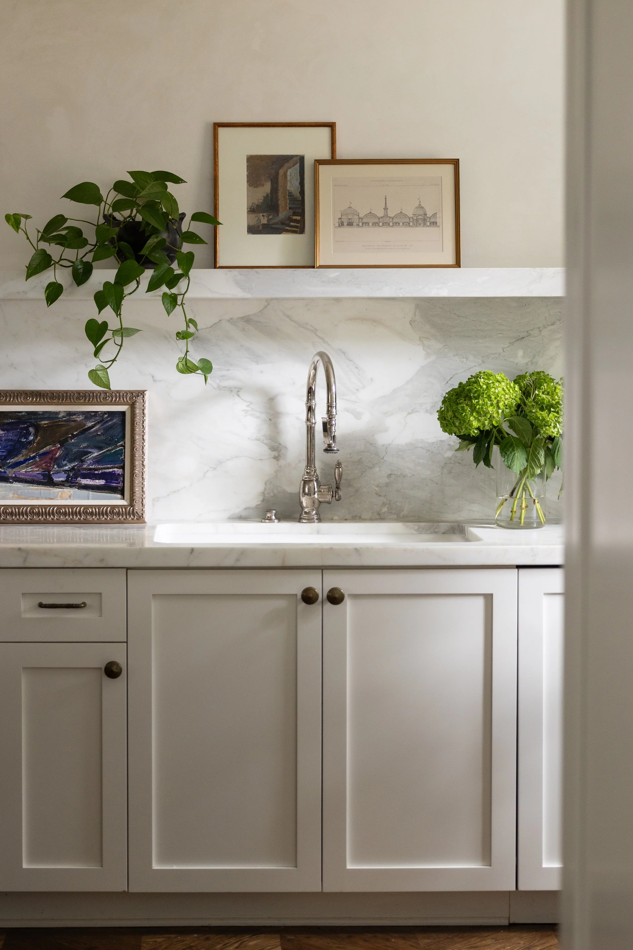 A kitchen sink with a white marble countertop, a brass faucet, a potted plant on the left side, framed artwork above, and a vase with green hydrangeas on the right side.