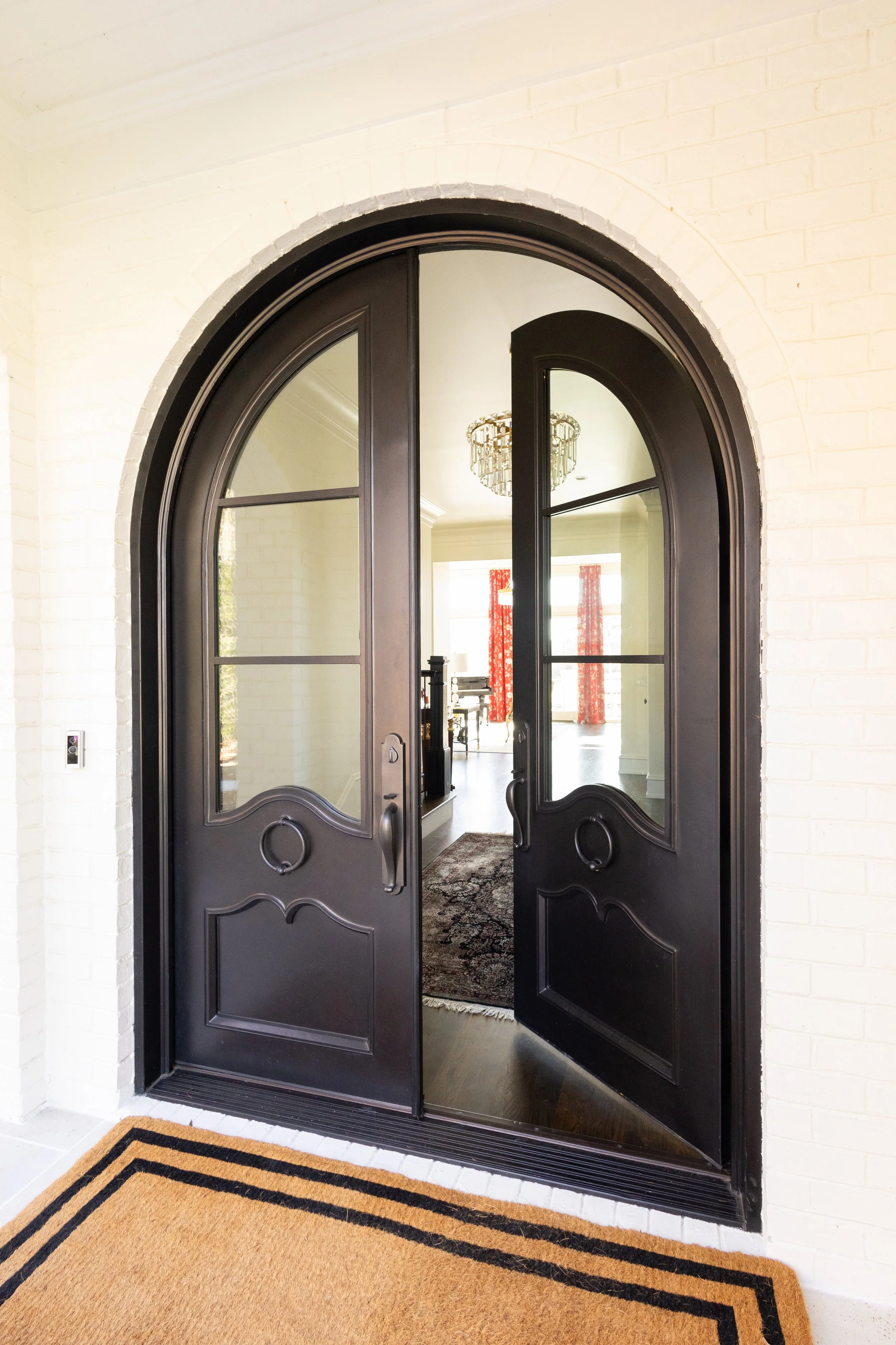 Open black arched double front door with glass panels, leading into a bright room with a chandelier, red curtains, and a patterned rug.