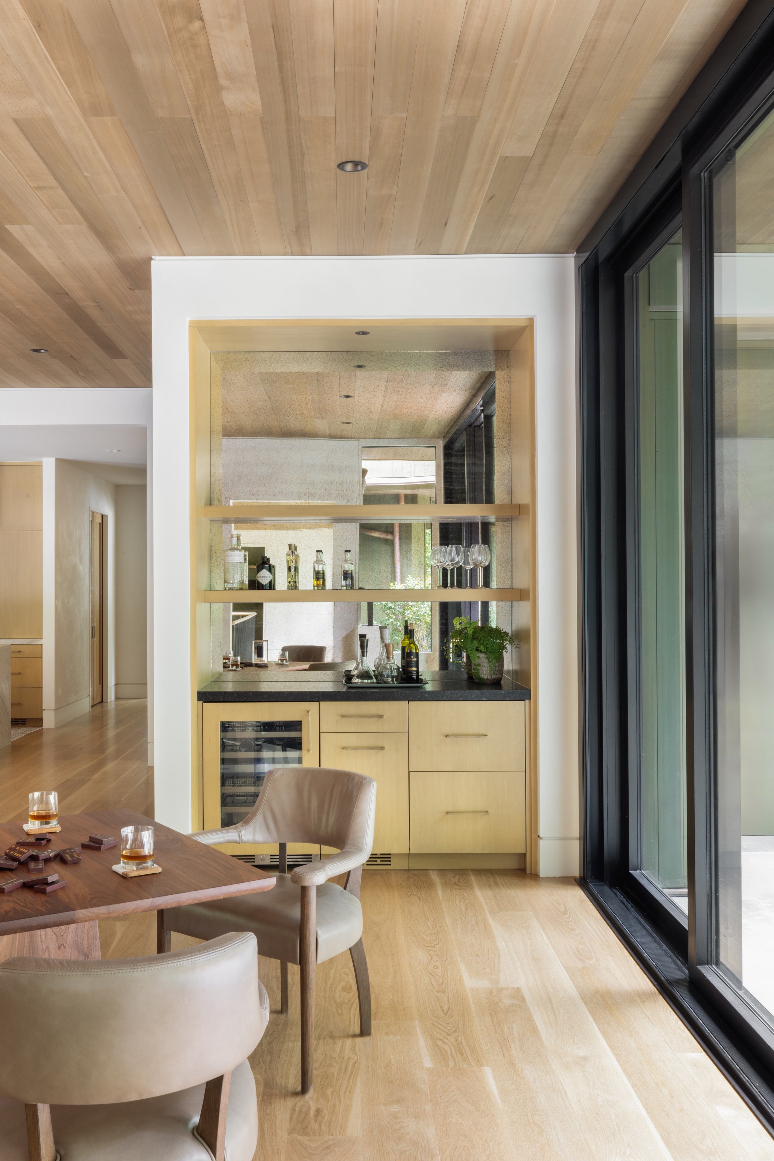 Modern dining room with a built-in bar area, wooden shelving, and large glass sliding doors leading outside. Light-colored wood flooring and ceiling, with beige chairs around a wooden dining table.