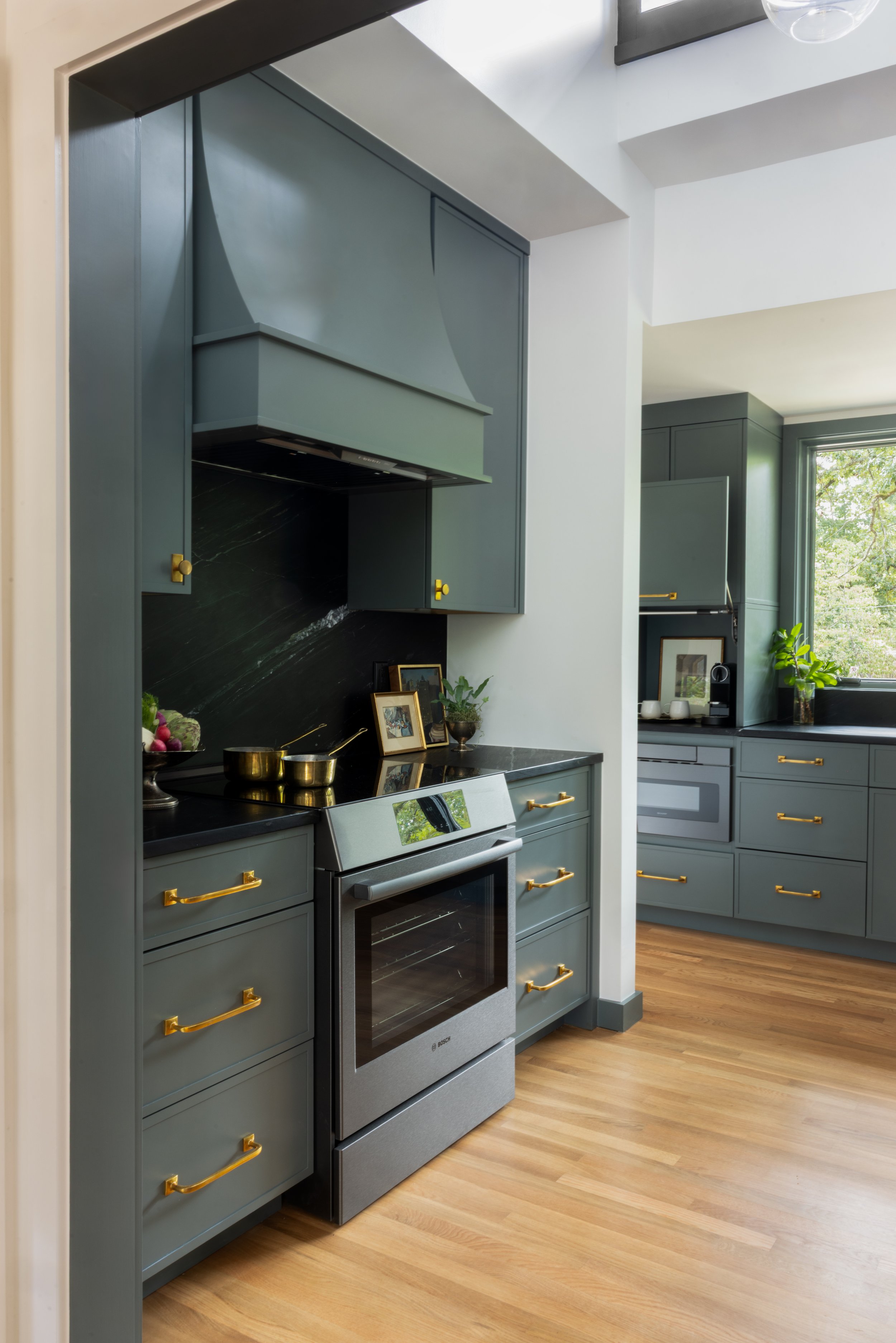 Modern kitchen with blue cabinetry, black countertops, a stainless steel oven, and gold handles, with hardwood floors and a window showing greenery outside.