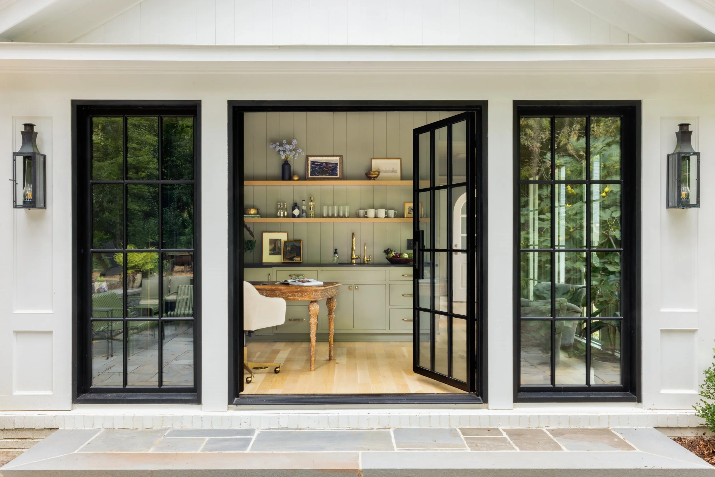 View of a modern kitchen seen through black-framed glass doors, with a dining area, light green cabinets, and open shelving inside.