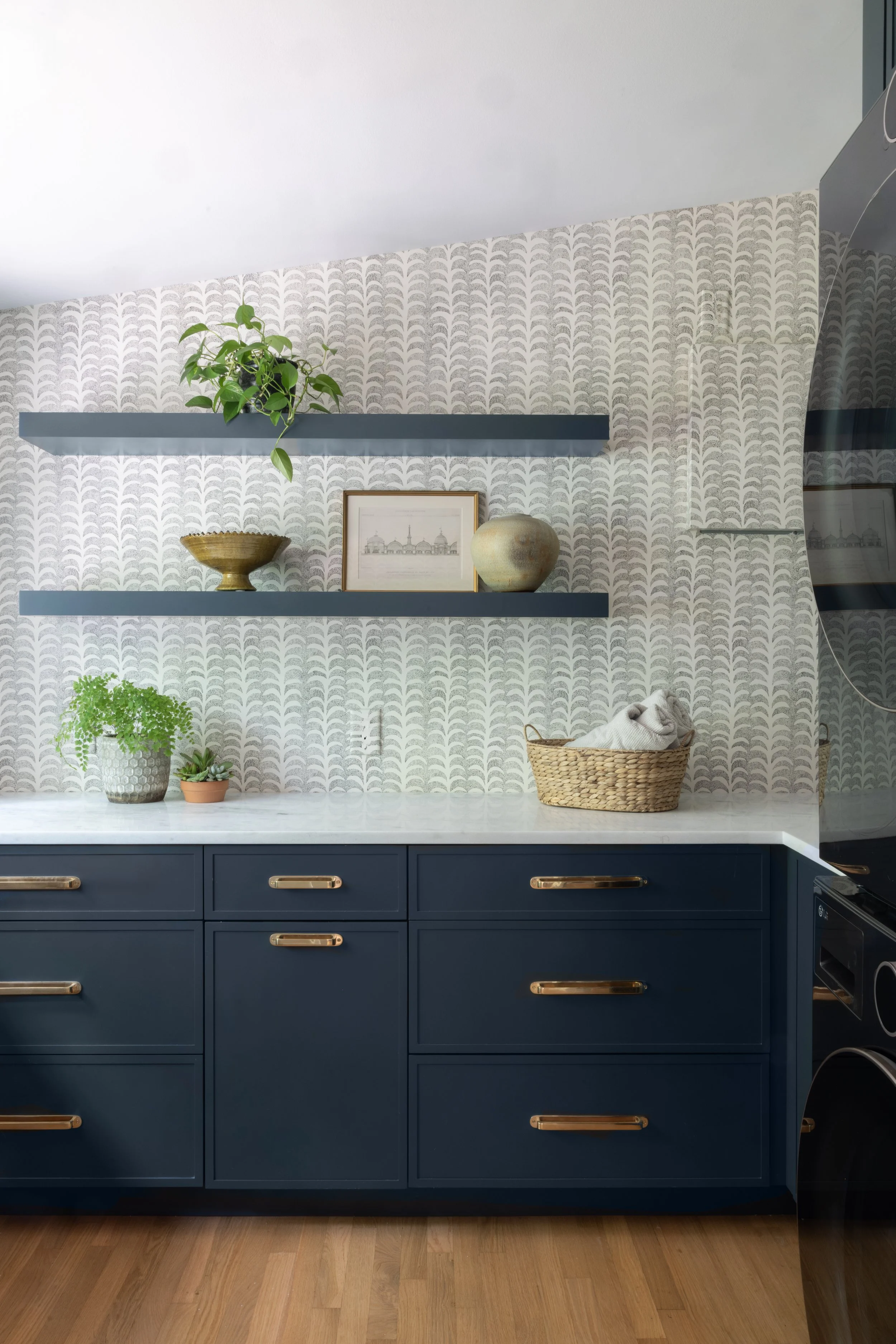 A modern kitchen with navy blue cabinets featuring gold handles, a white marble countertop, decorative objects on open shelves, and patterned wallpaper.