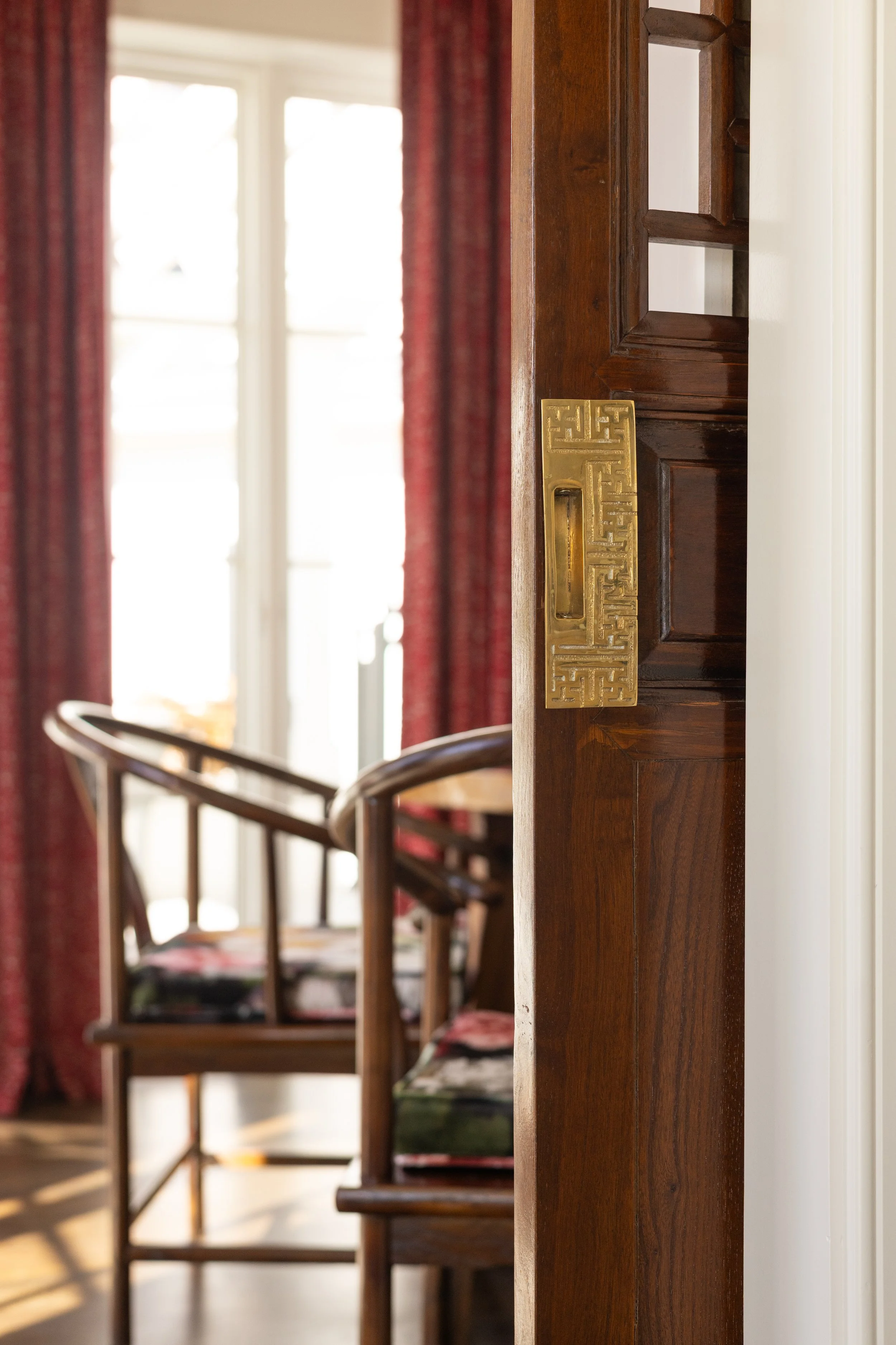 Partially opened wooden door with brass hardware leading to a room with two wooden chairs and large windows with red curtains letting in bright sunlight.