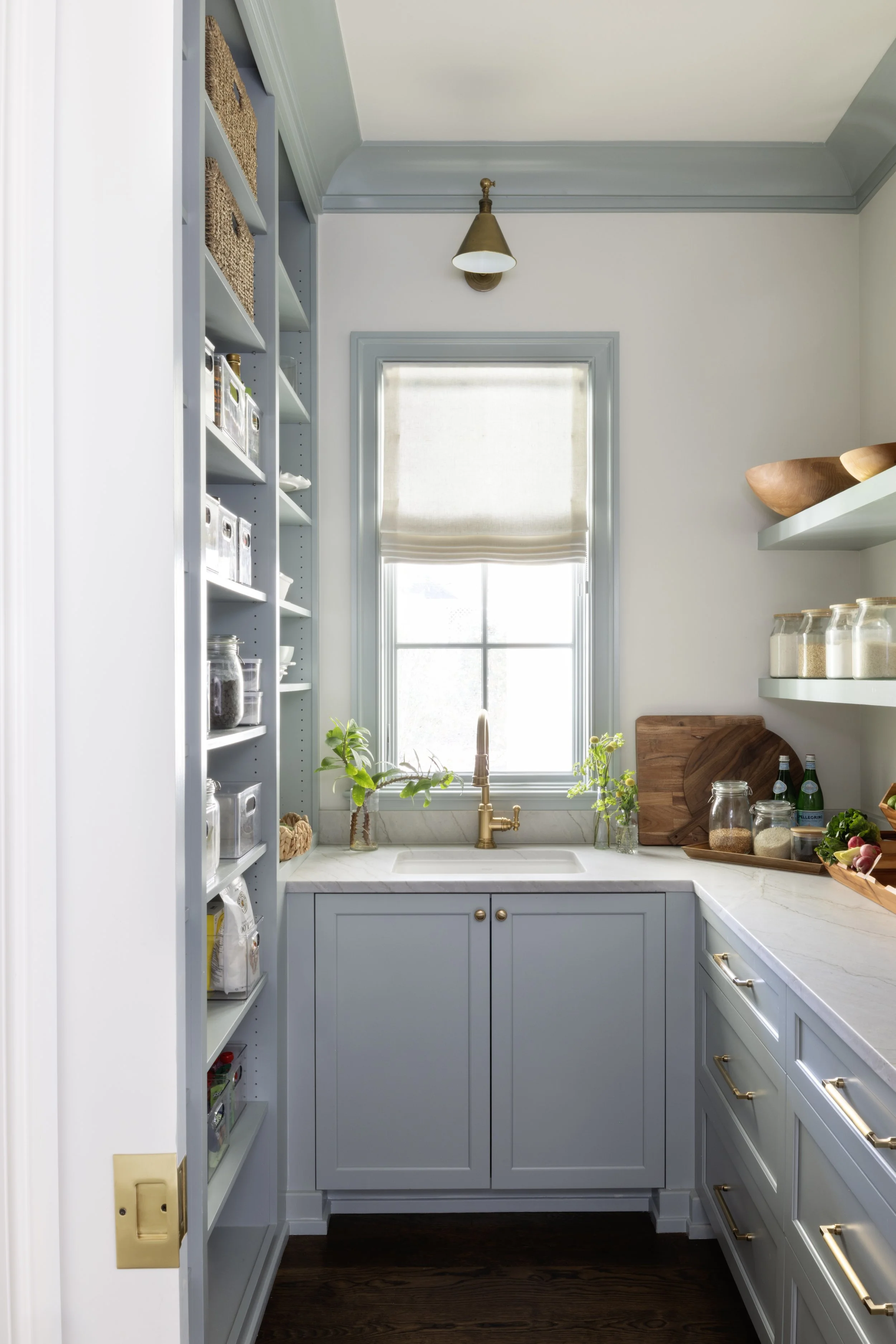 A small kitchen with light blue cabinetry, a marble countertop, and a single window with a beige Roman shade. There are glass jars on shelves, a wooden cutting board, green plants on the counter, and a brass faucet above the sink.
