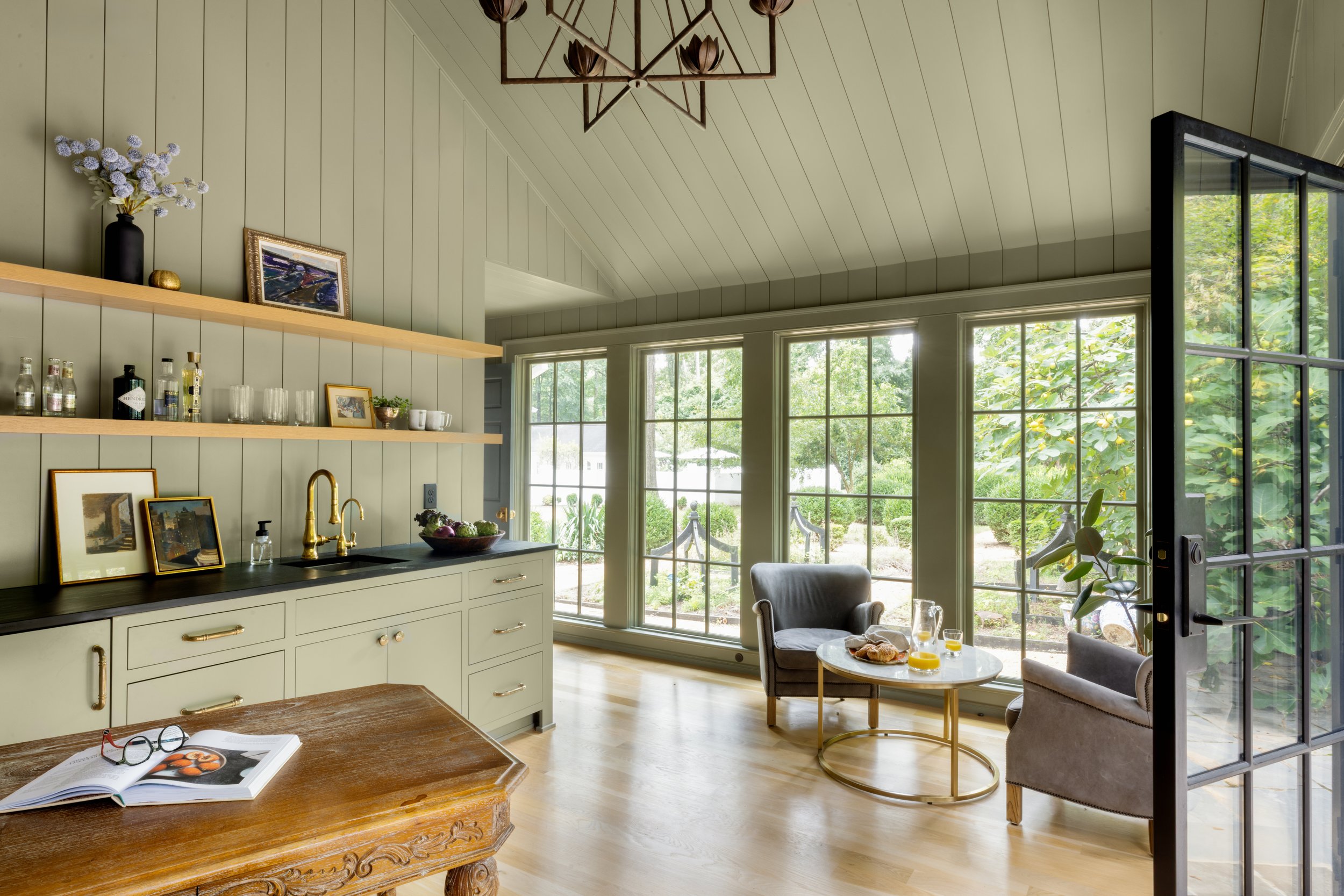 Cozy kitchen and sitting area with large windows and greenery outside, featuring a bathroom sink, open shelving, and a table with breakfast items.