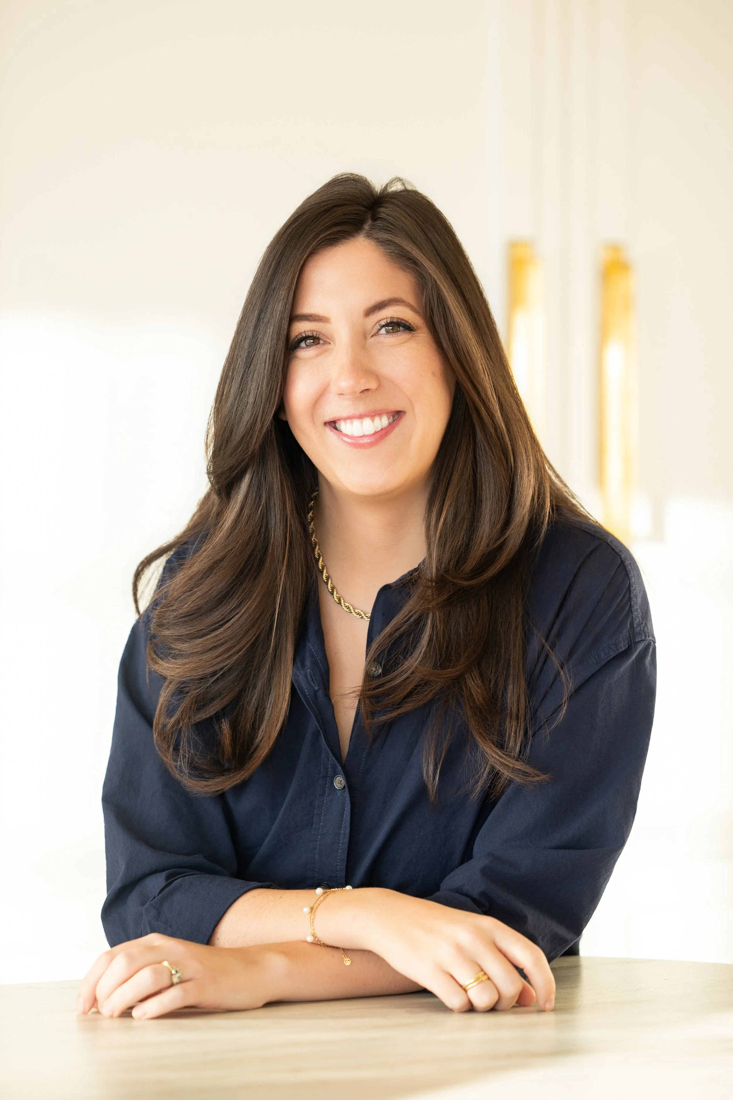Woman in floral dress sitting at a wooden table with papers, pens, and a tumbler in a bright room with large windows.