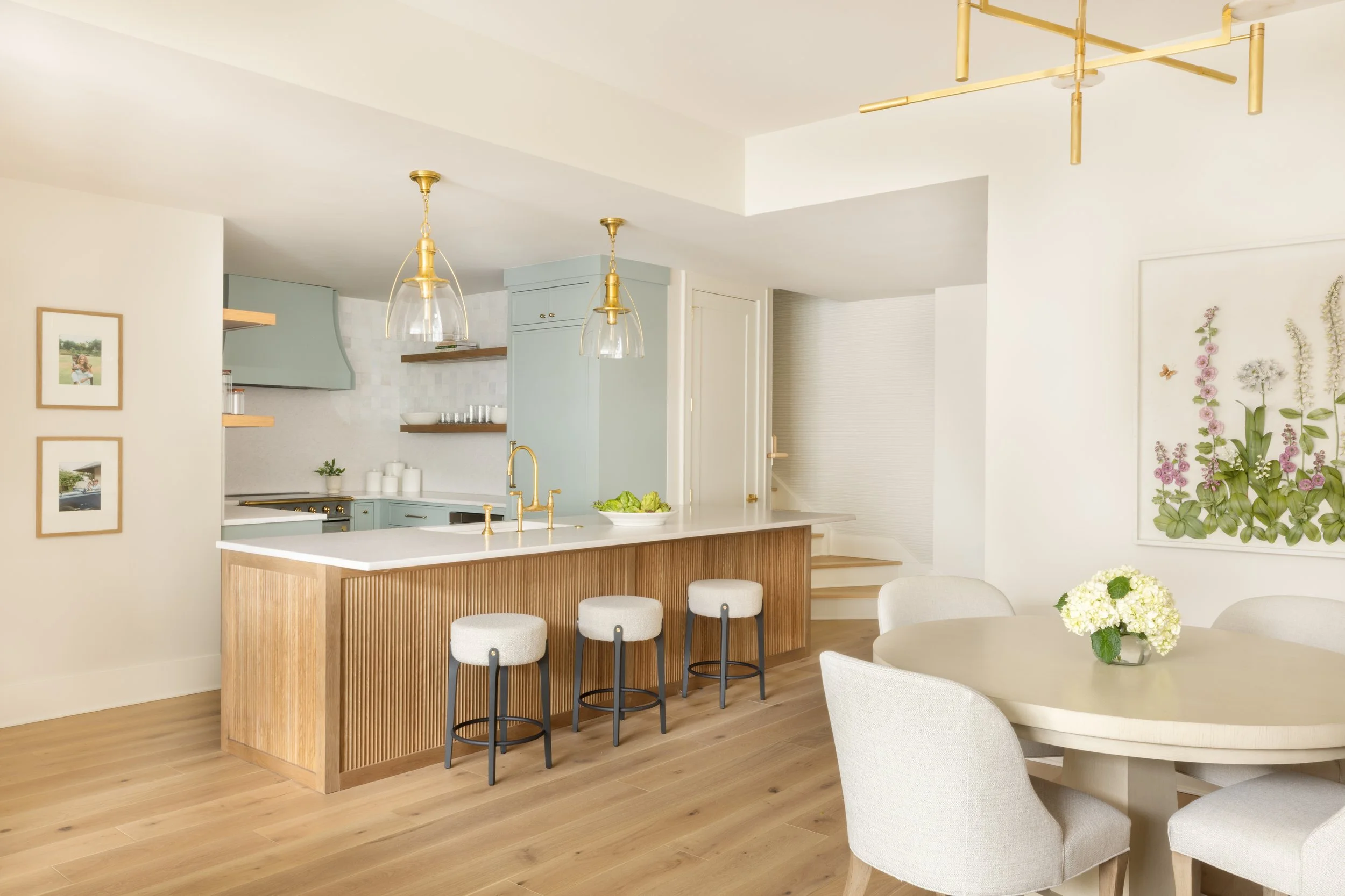 Bright kitchen and dining area with light wood flooring, white walls, and minimalistic furniture. The kitchen has pastel green cabinets, a white countertop, and gold fixtures, including a faucet and pendant lights. There is a round dining table with 