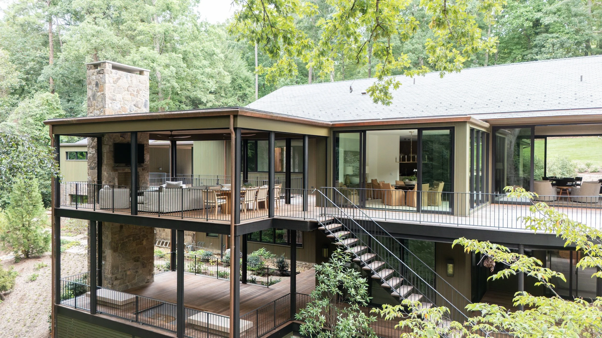 Multi-level modern house with large outdoor decks, glass sliding doors, and outdoor furniture situated in a lush, green wooded area.