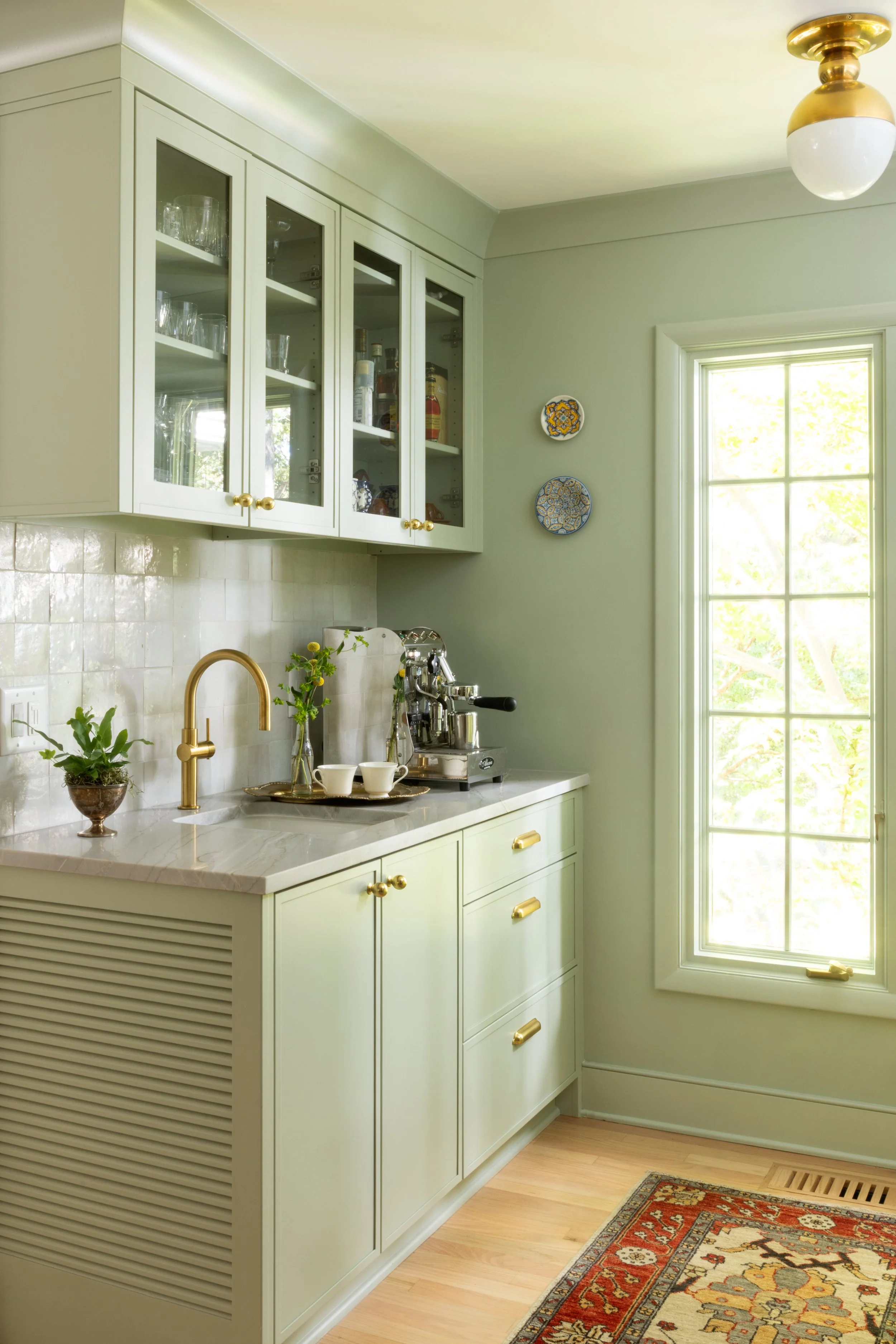 A light green kitchen with a window, a gold faucet, white countertop, glass-front cabinets, a coffee machine, and decorative plates on the wall.