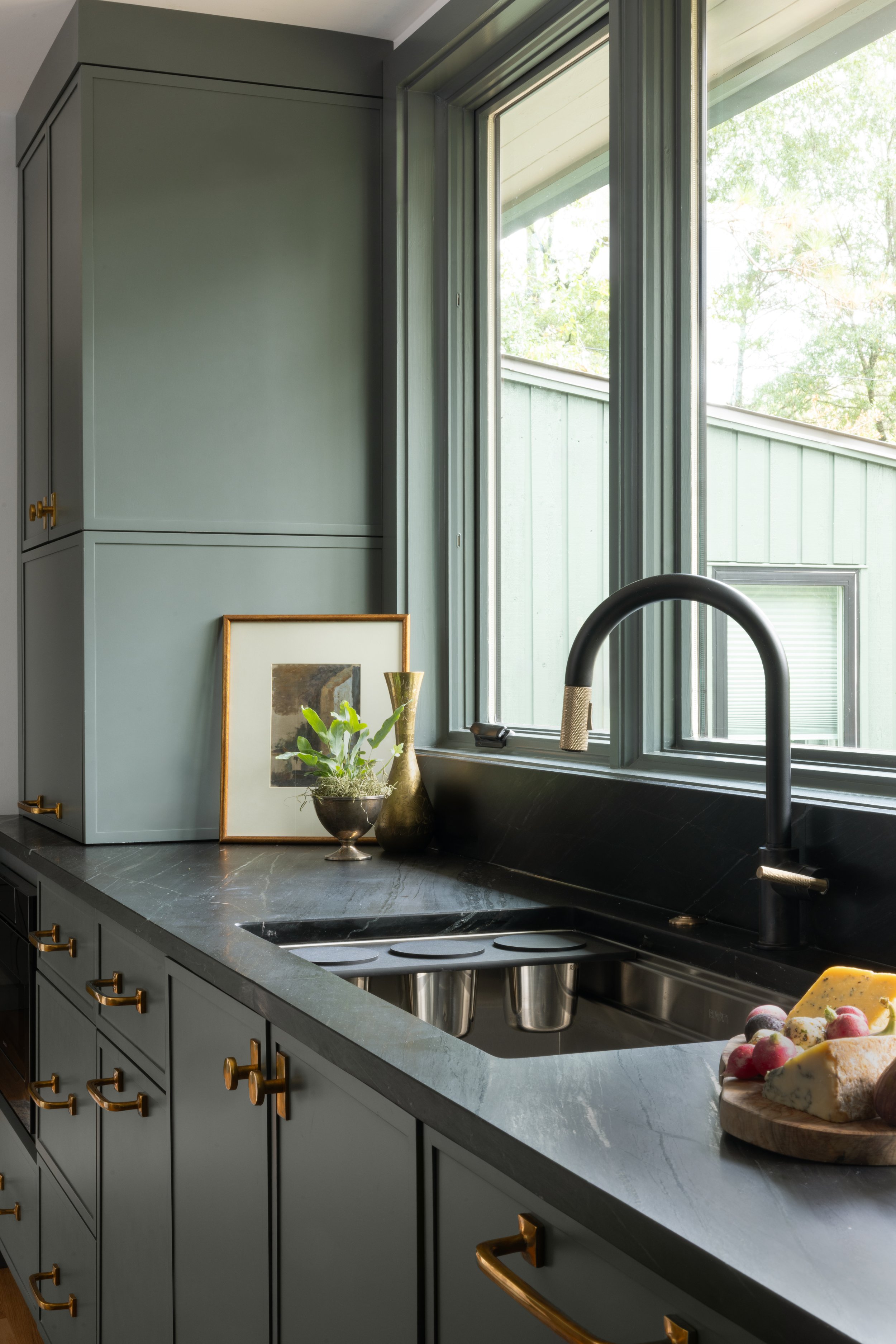 Kitchen with green cabinets, black countertop, black sink, and large window with greenery outside.