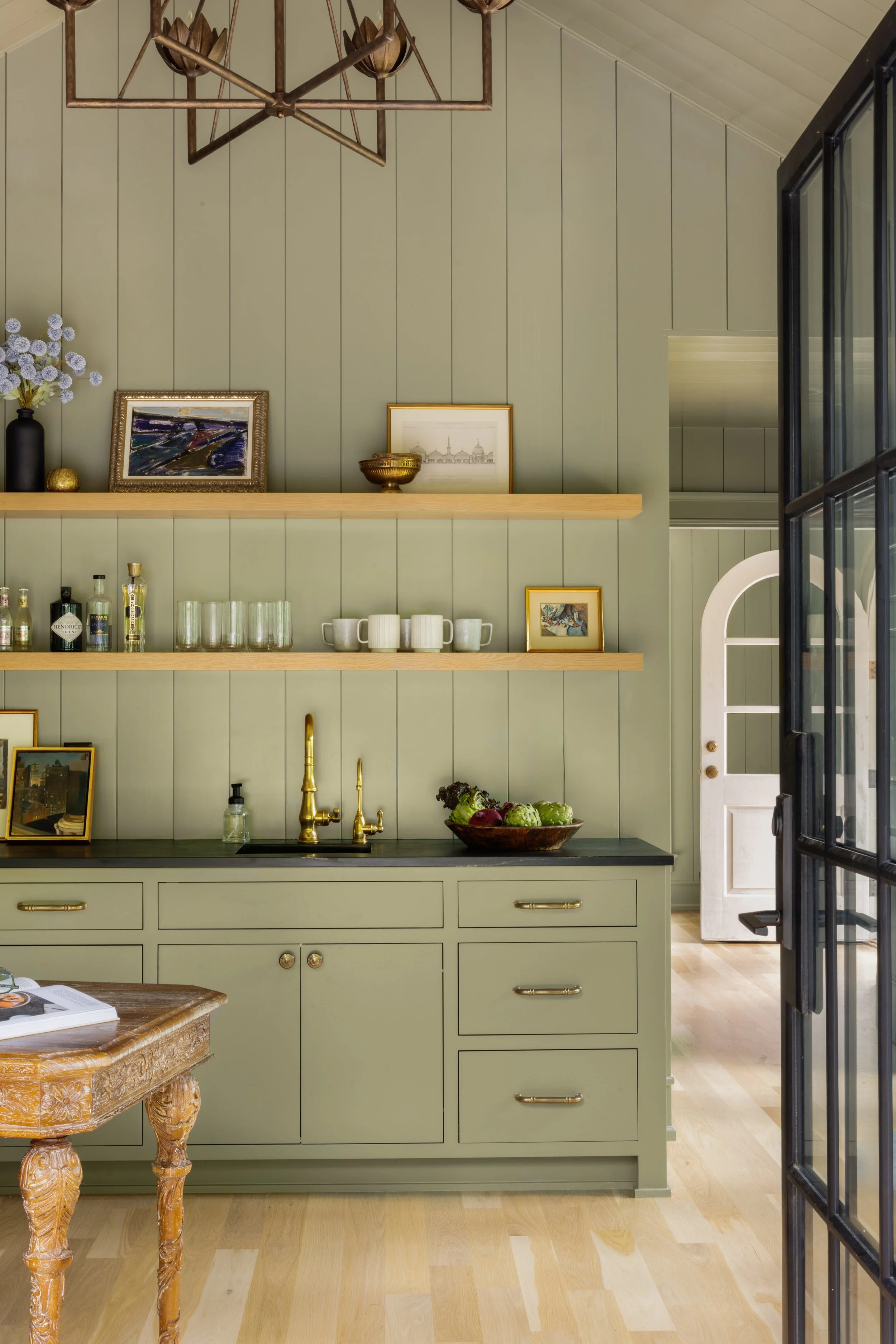 A kitchen with sage green cabinets and open wooden shelves displaying decorative items, glasses, and framed artwork. There is a black countertop with a brass faucet, a bowl of vegetables, and a white door with an arched window in the background. Part
