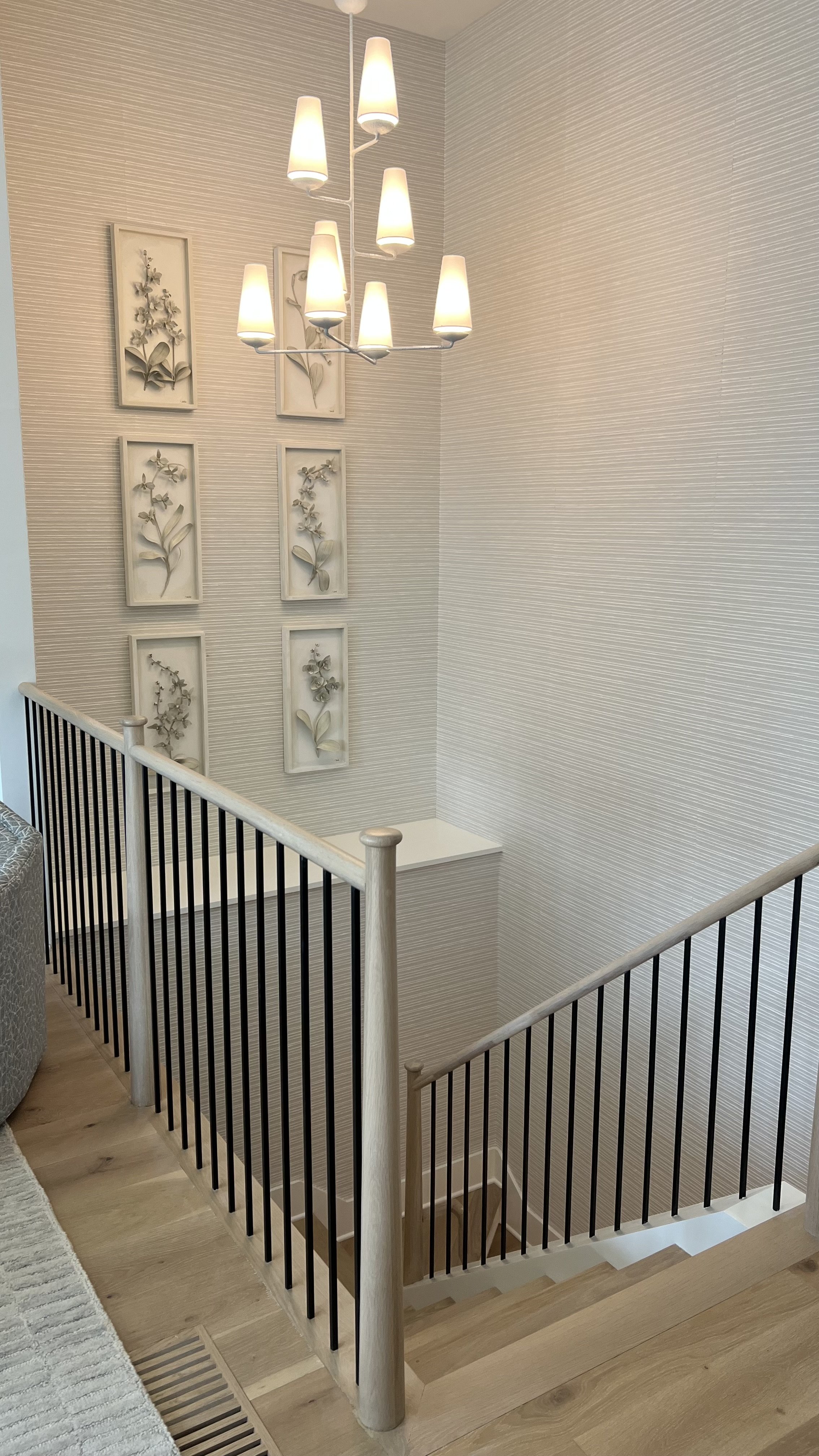 A staircase with a wooden handrail and black balusters, leading down to a lower floor. The upper landing has a white wall with textured wallpaper, decorated with seven framed pressed plant specimens arranged vertically. A modern chandelier with multi