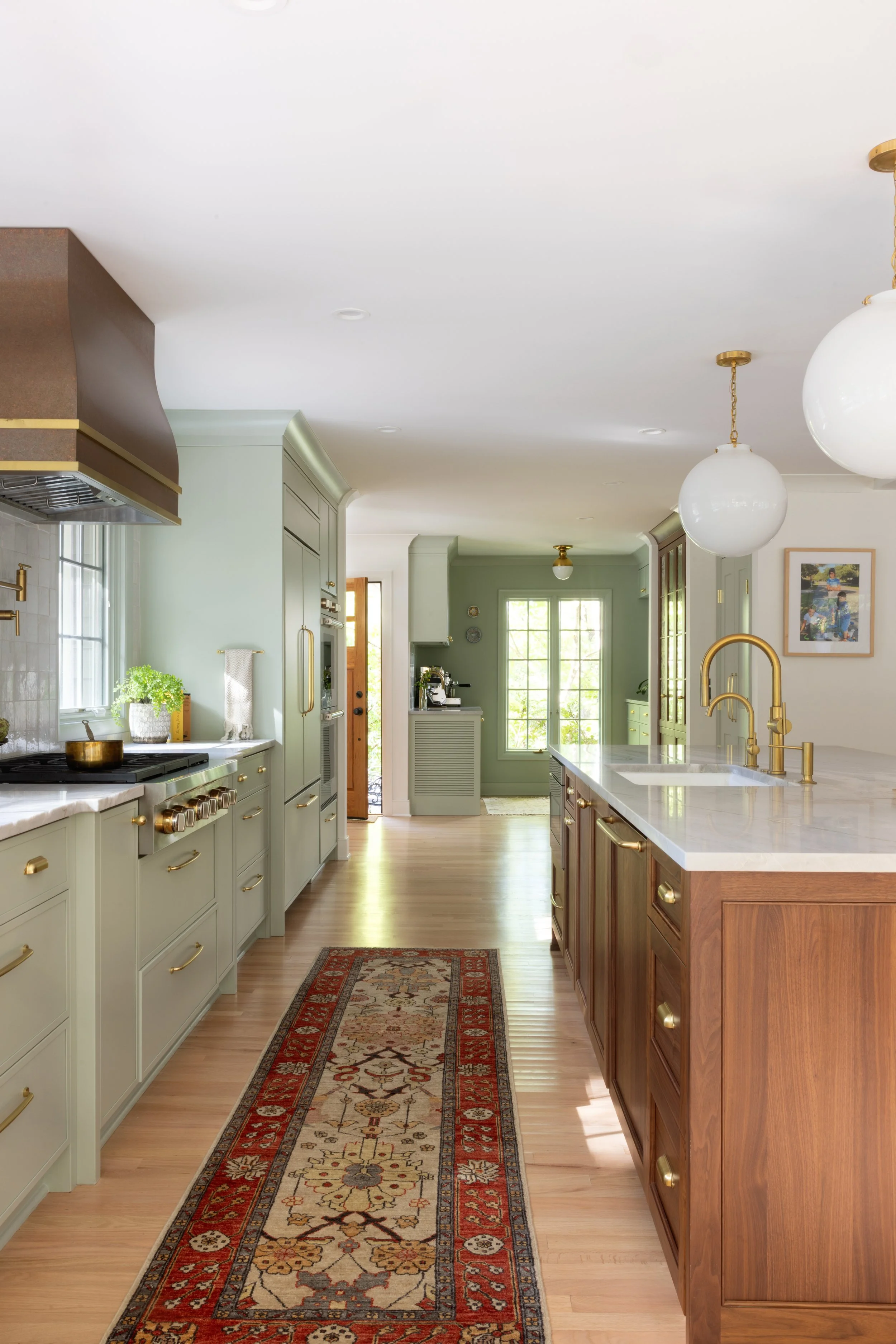 Bright kitchen with light green cabinets, a wooden kitchen island with brass hardware, white marble countertops, and gold fixtures. There is a patterned rug on the hardwood floor, two large white globe pendant lights hanging from the ceiling, and a w
