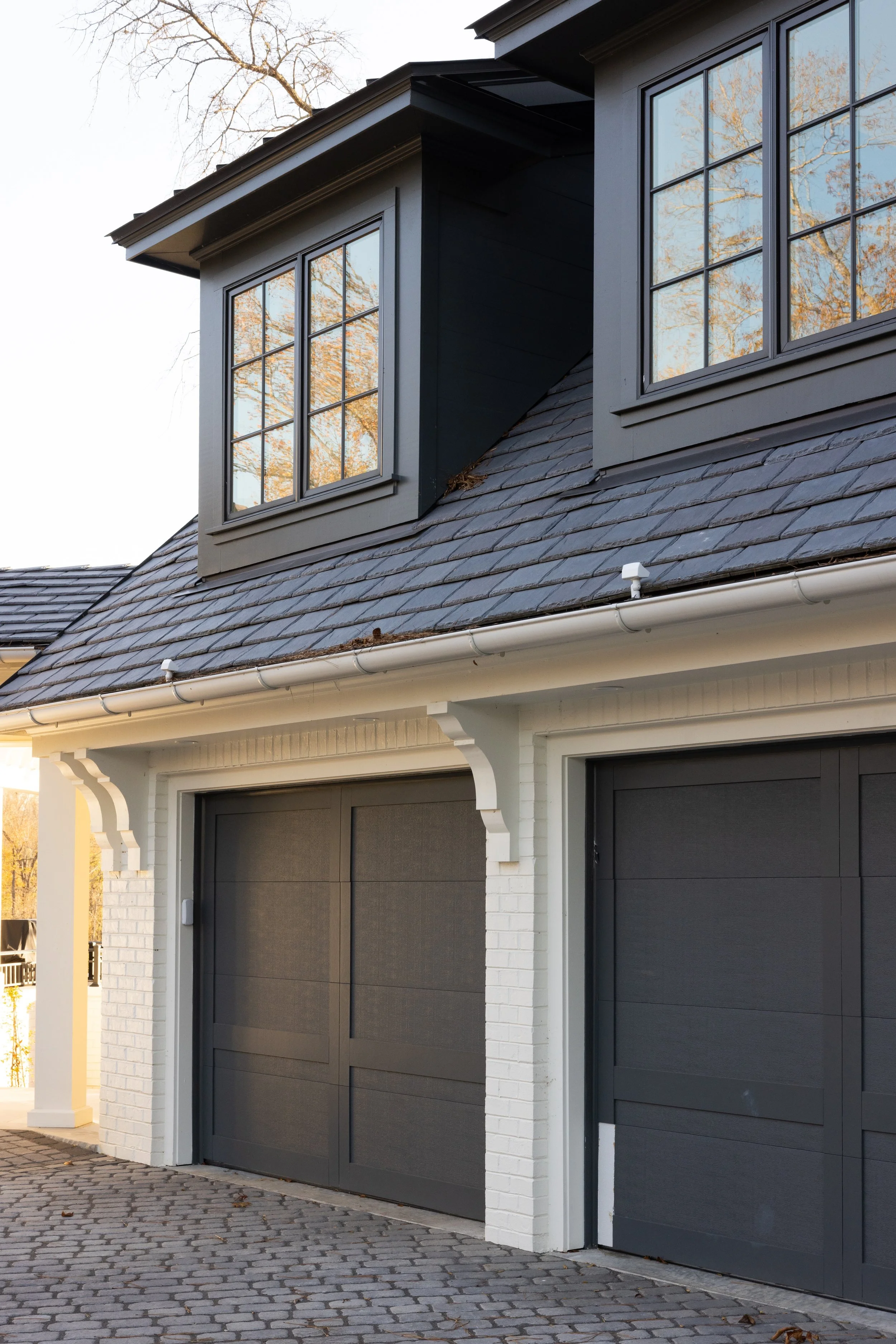 Exterior view of a house with dark garage doors, white brick pillars, gray window dormers, and a slate roof, with leafless trees reflected in the window panes.