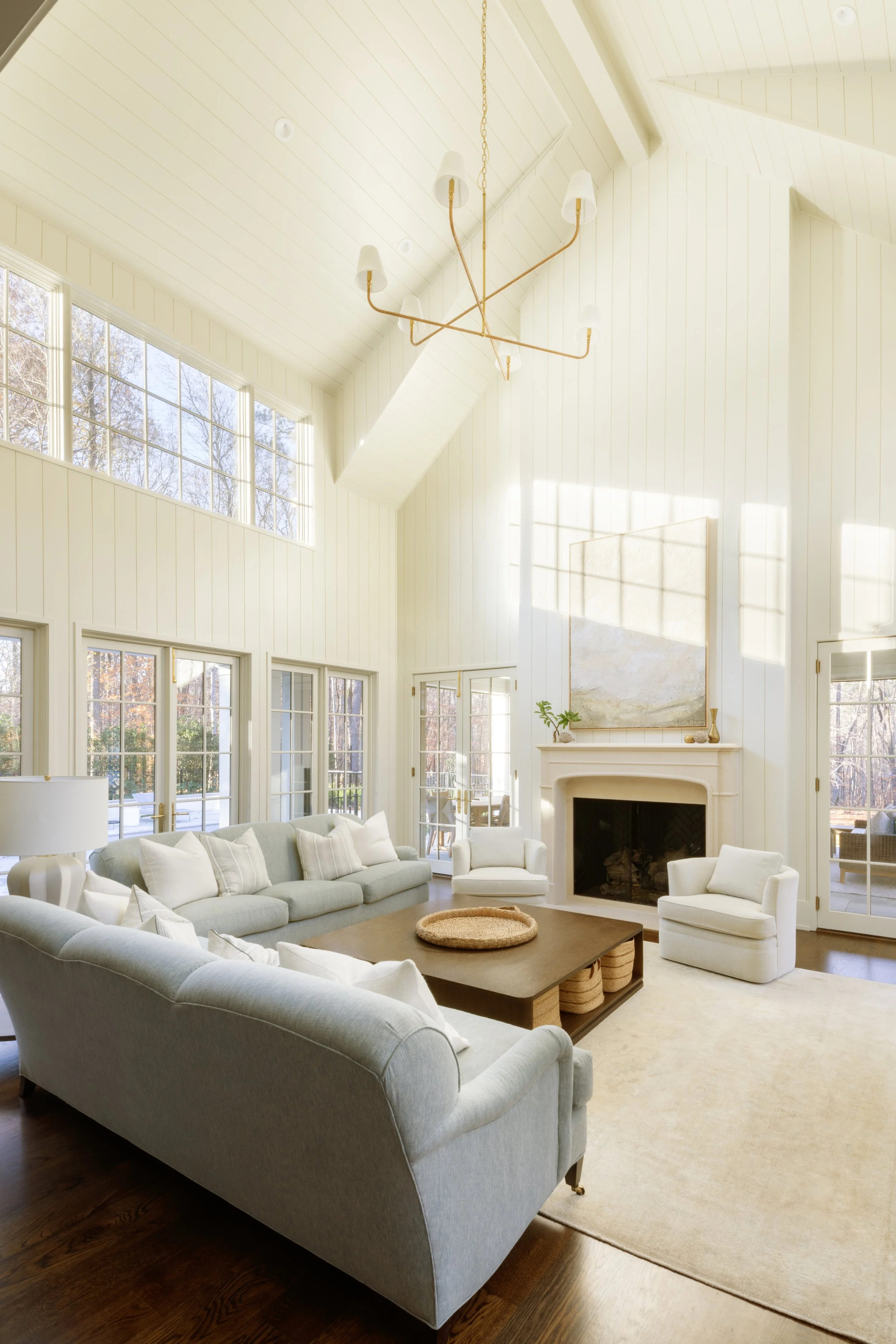 Bright living room with large windows, white walls, a fireplace, and white furniture including sofas and armchairs, with a wooden coffee table and a beige rug.
