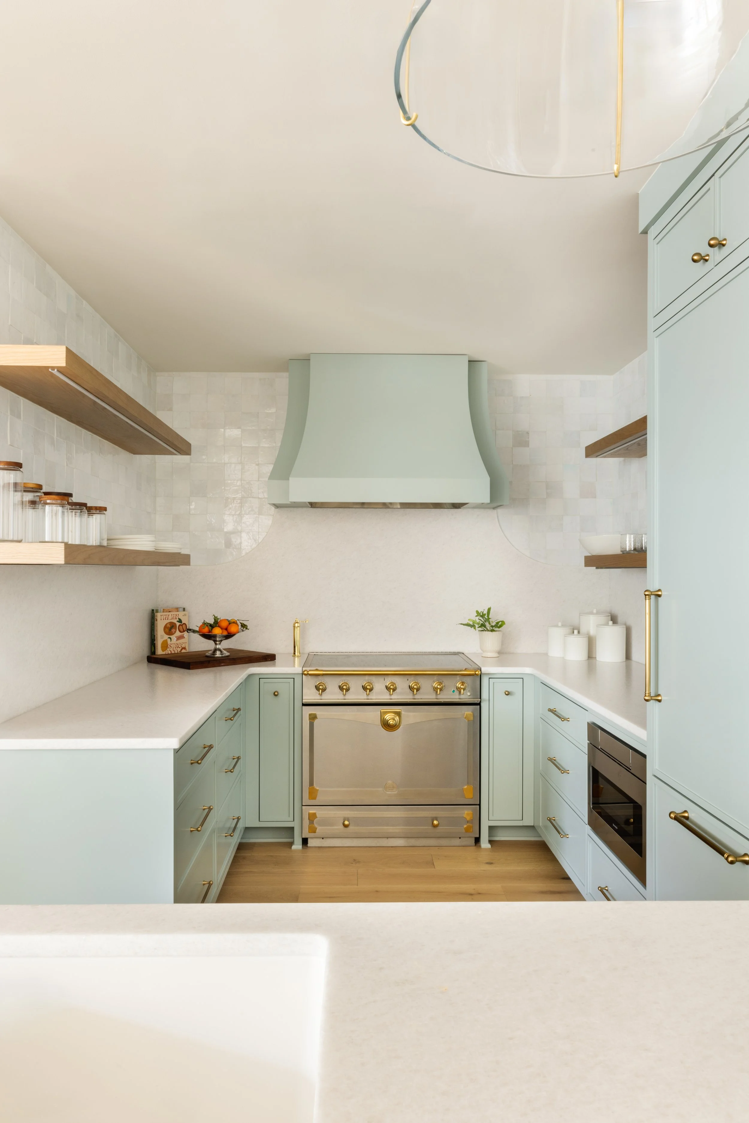 A modern kitchen with light blue cabinetry, gold hardware, a stainless steel oven, and open wooden shelves with glass jars and white dishes. A white range hood is centered above the stove, and the countertops hold a fruit bowl, a book, and a small pl