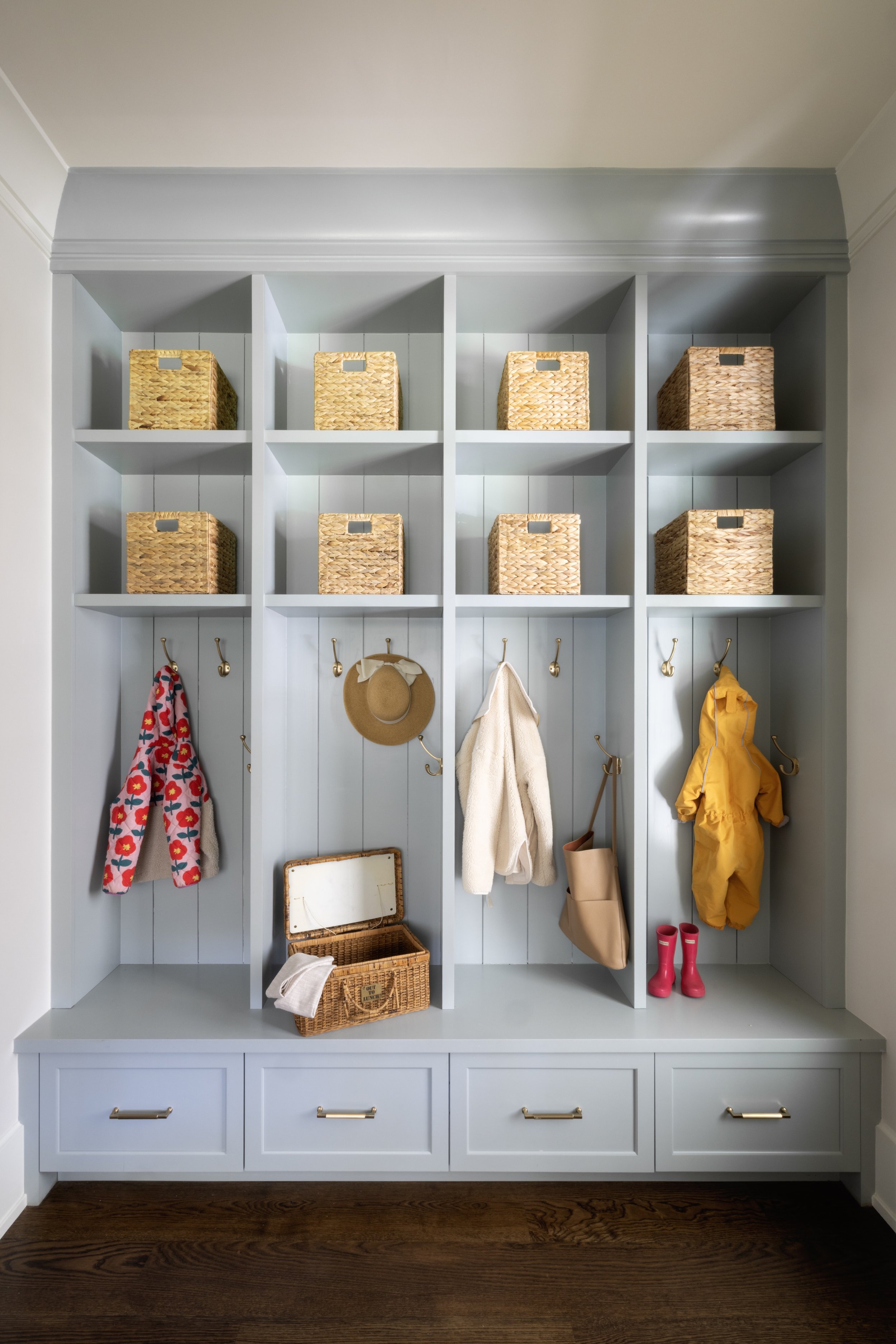 Entryway cubby storage unit with wicker baskets, hooks with coats and hats, and pink rain boots on a hardwood floor.