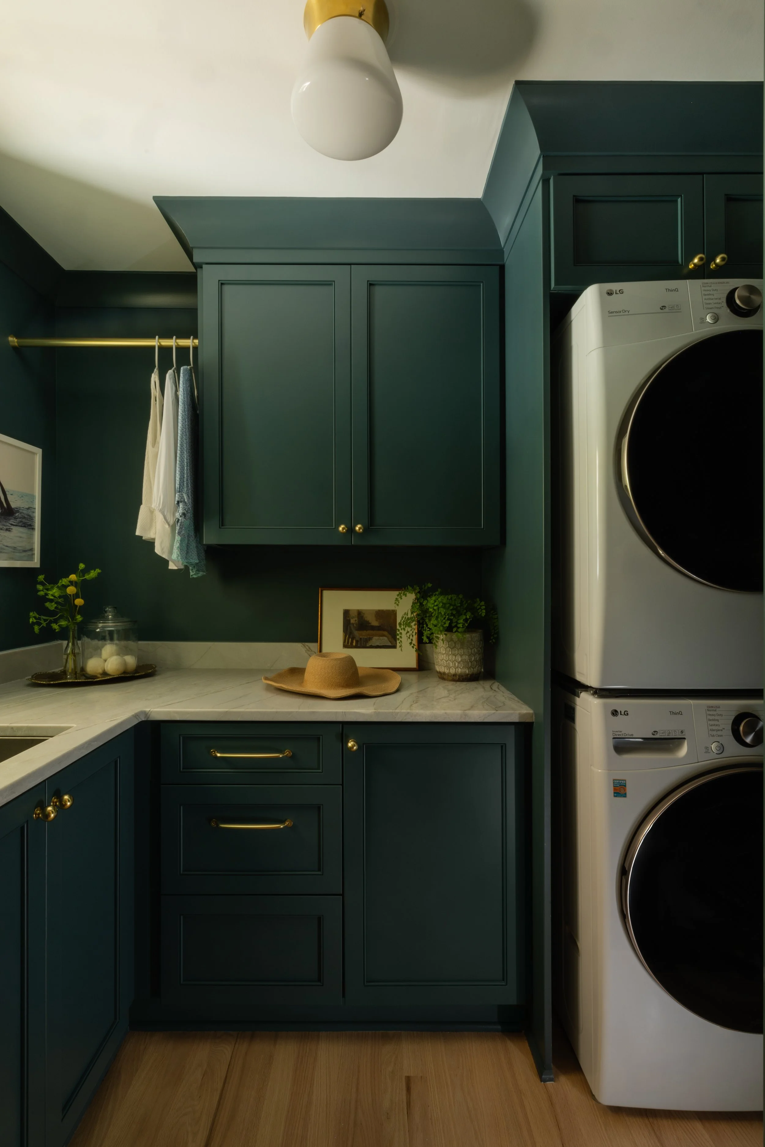 Laundry room with dark green cabinets, laundry washer and dryer, marble countertop, hanging clothes, and decorative items.