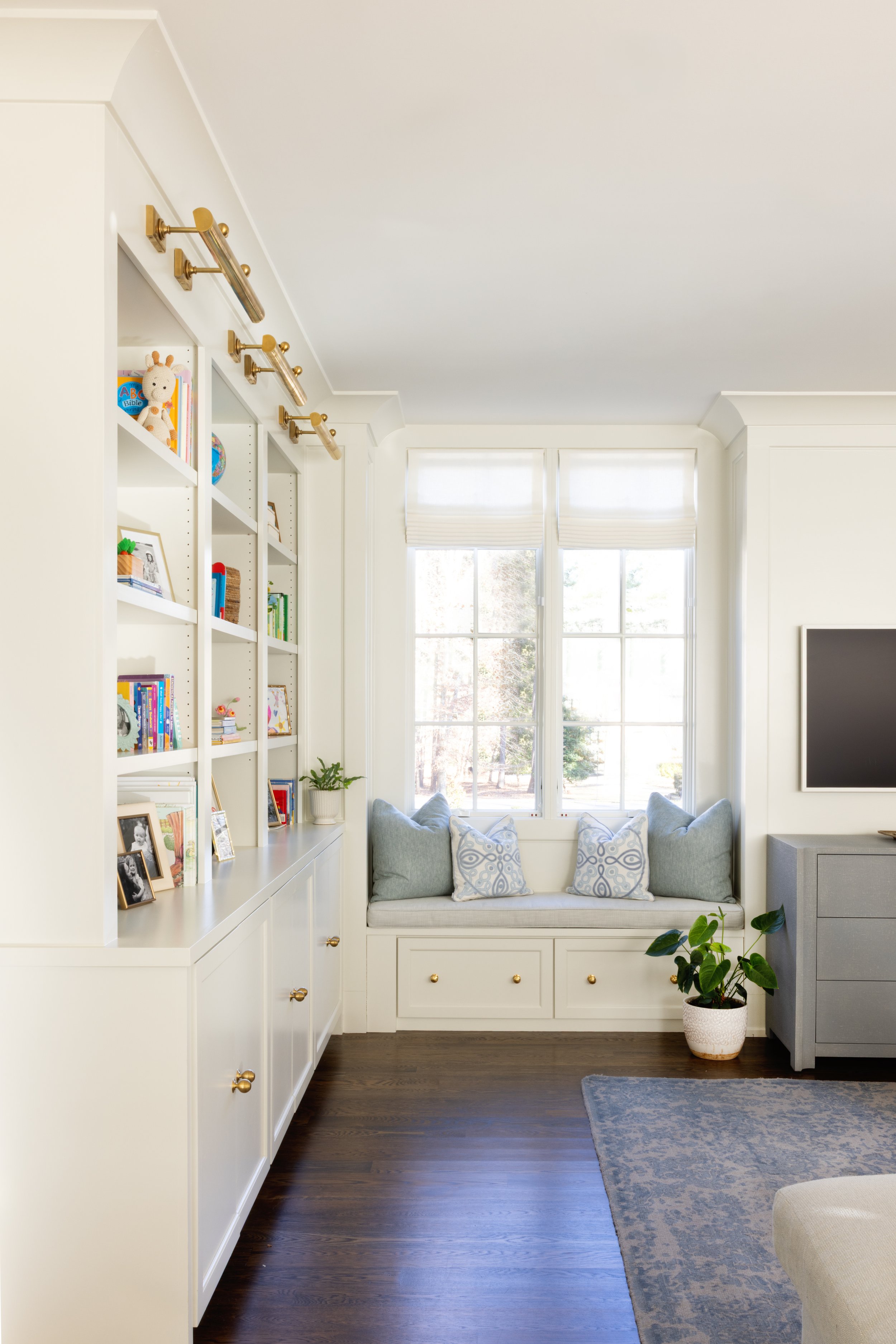 Sunlit living room with white built-in bookshelf and window seat, decorated with cushions and potted plant, dark hardwood flooring, and grey dresser with mounted TV.