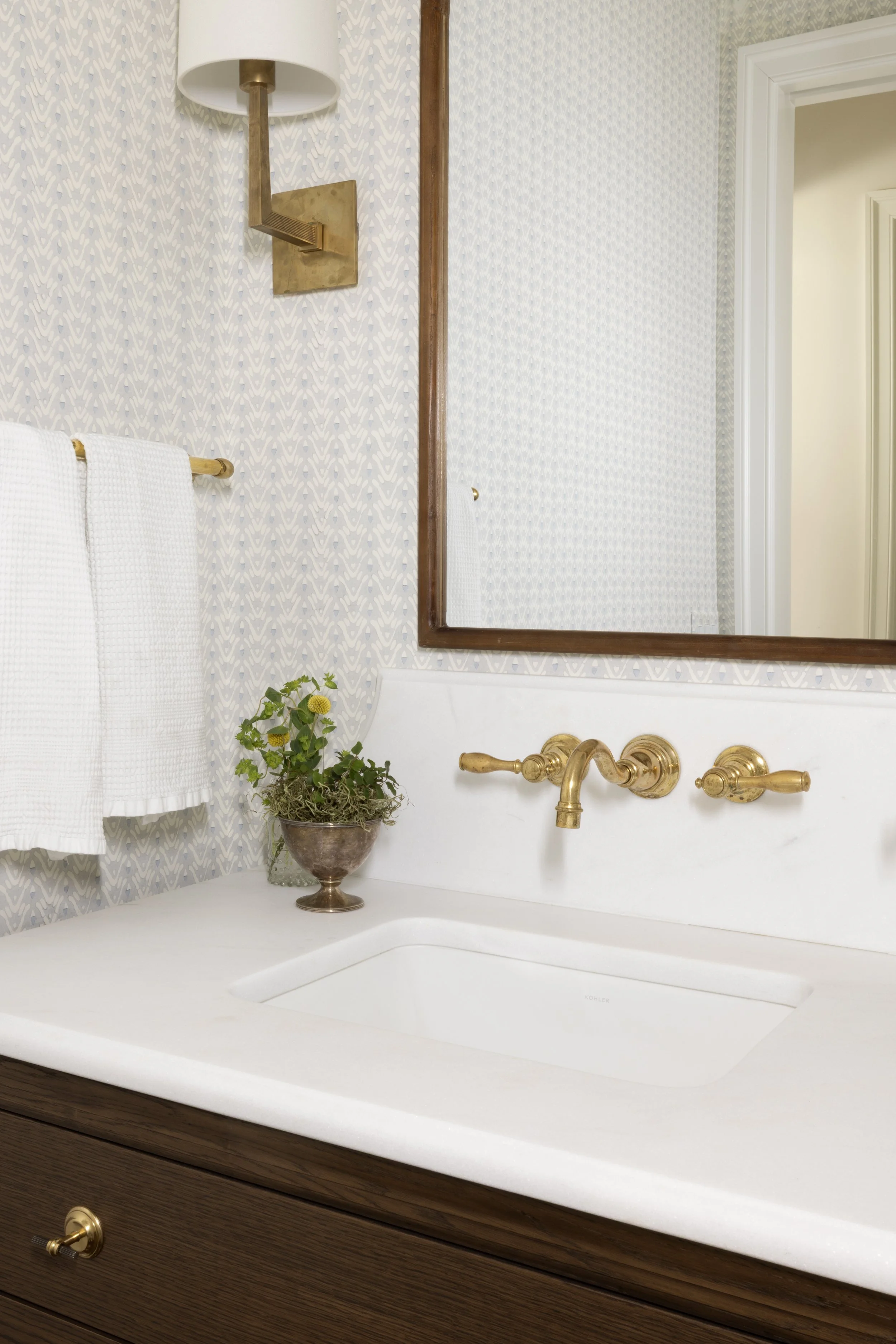 Bathroom sink with gold fixtures, mirror, white towels, a small plant in a metallic pot, and Wallpaper with a pale blue pattern.