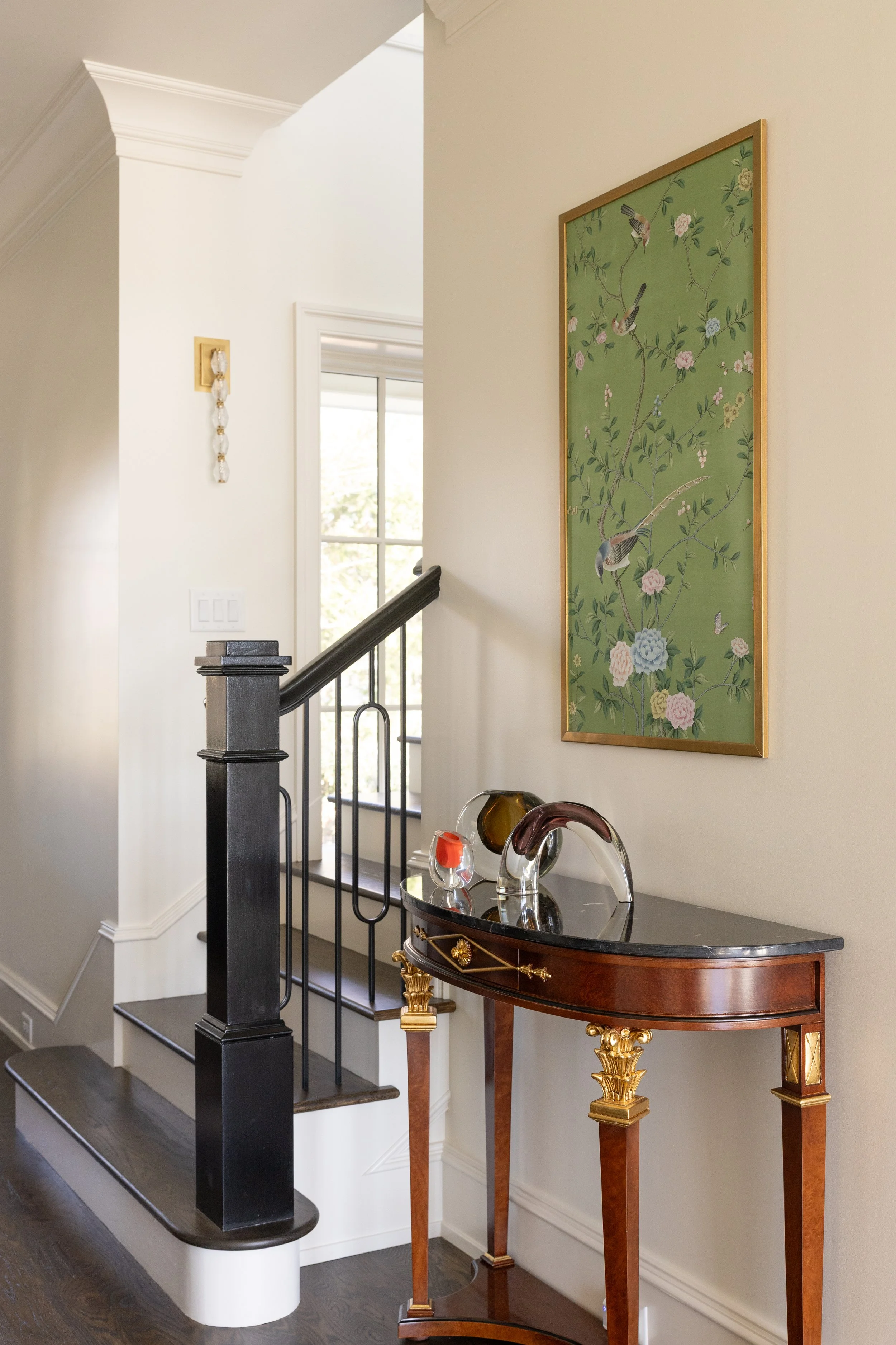 A hallway corner with a dark wooden console table with gold accents, a black marble top, and decorative glass sculptures on it. Above, a large green framed artwork featuring birds and flowers hangs on the wall. A staircase with black railings leads u
