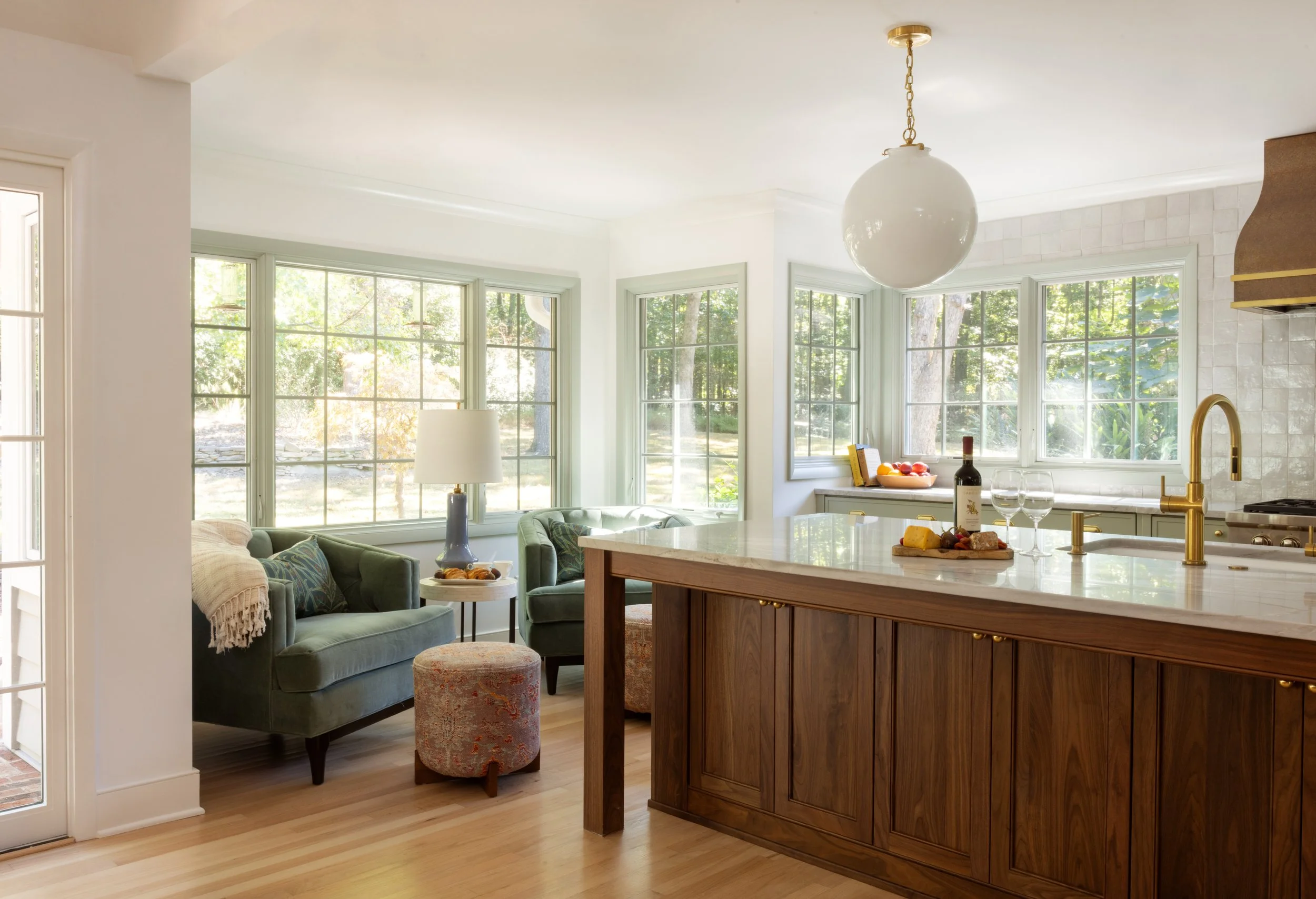 Bright kitchen and living room with large windows, green sofa, table lamp, wooden kitchen island, and outdoor greenery visible through the windows.