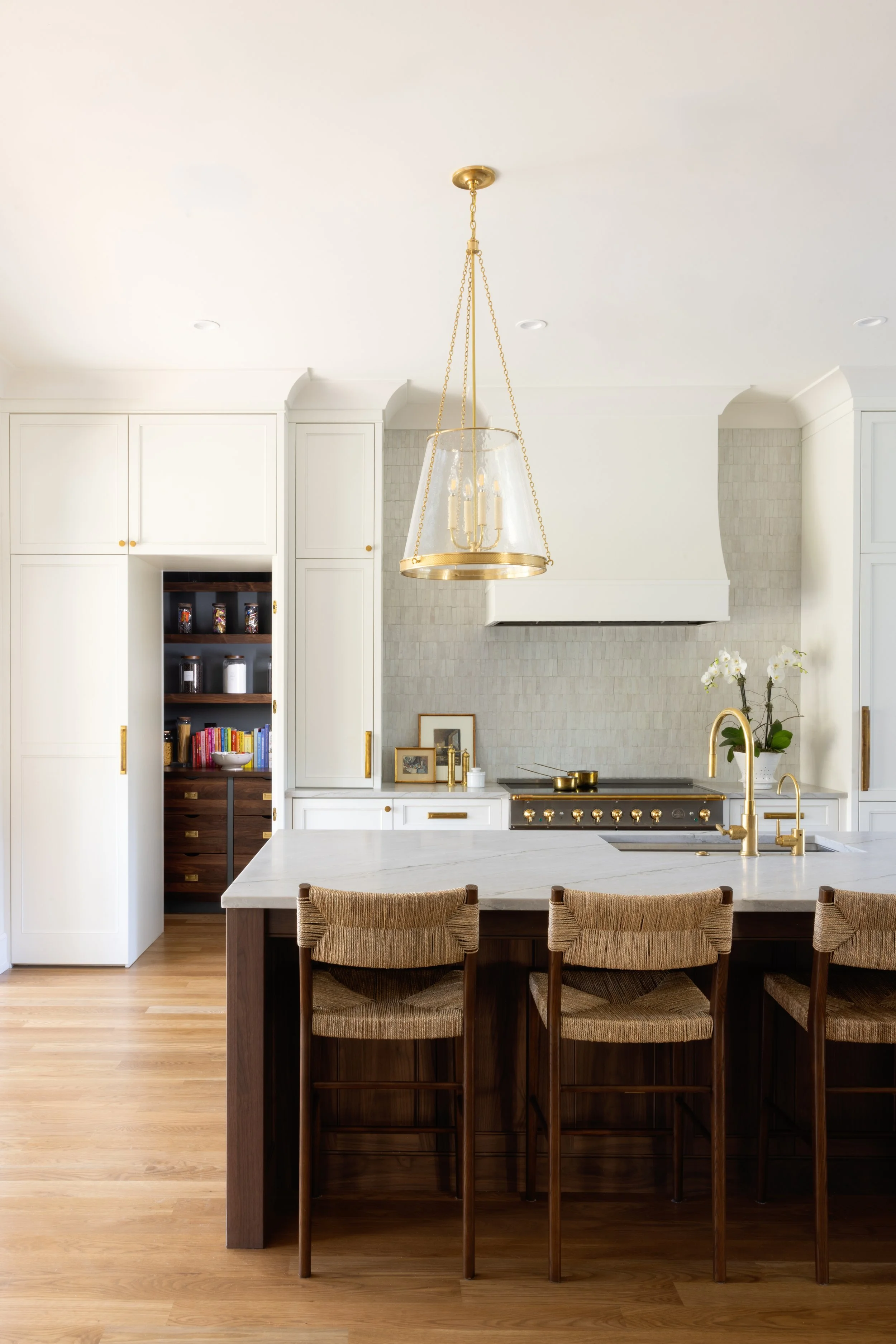 Modern kitchen with white cabinetry, a large island with a marble countertop, wooden bar stools, and gold fixtures. There is a hanging gold and glass chandelier, a stove with gold knobs, and a potted white orchid on the counter.,