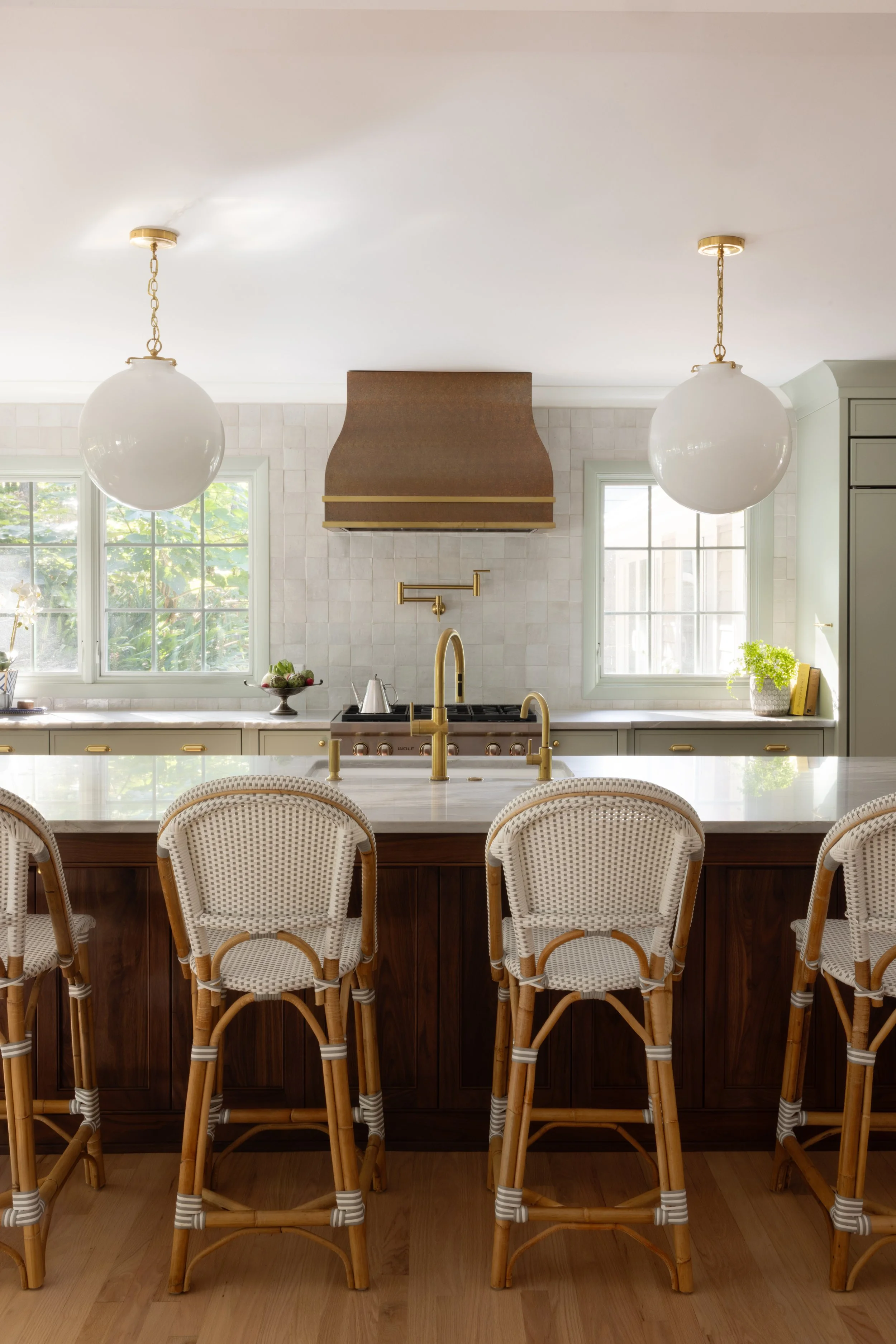 Bright kitchen with two large white pendant lights hanging from the ceiling, a white marble island with gold fixtures, brown wicker chairs with white woven seats, a vintage copper and brass range hood, white tiled walls, and green-framed windows lett