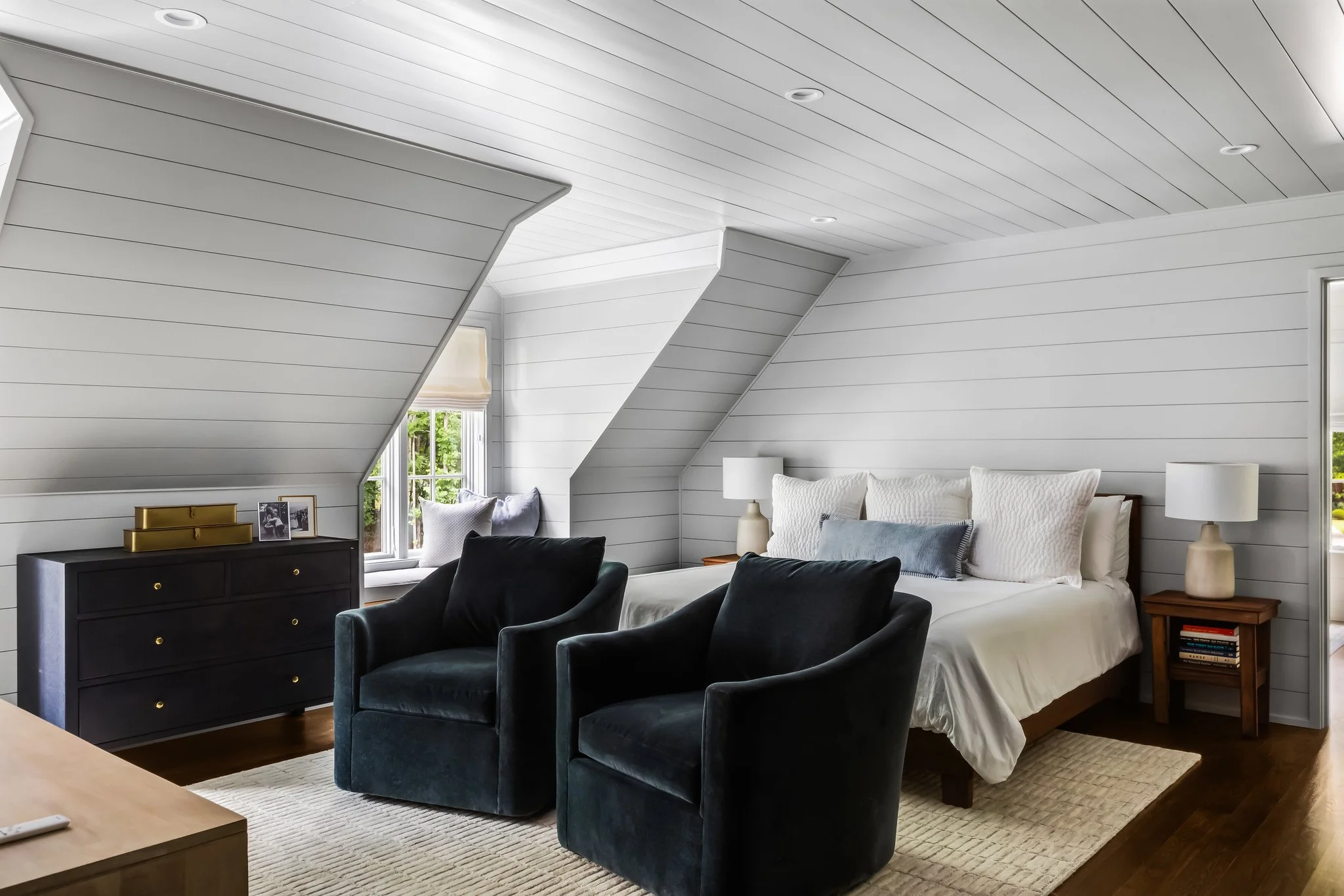 A cozy bedroom with a white bed, two side tables with lamps, two black armchairs, a dresser, and a window with daylight.