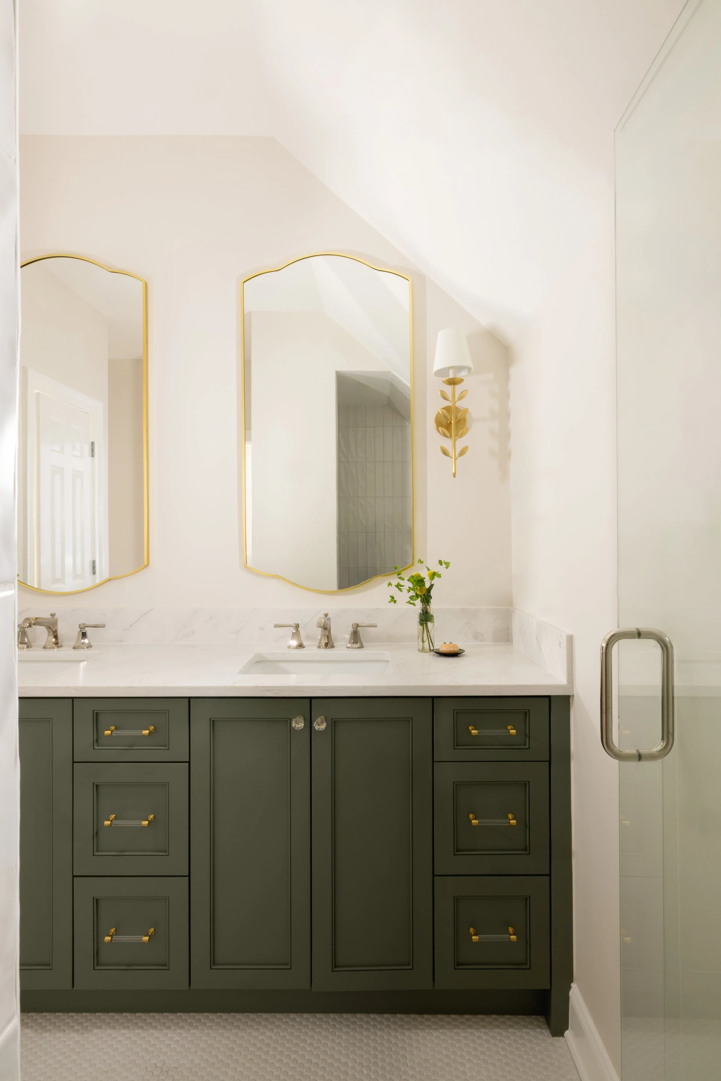 Bathroom vanity with two mirrors, green cabinets with gold handles, marble countertop, and a vase with yellow flowers.