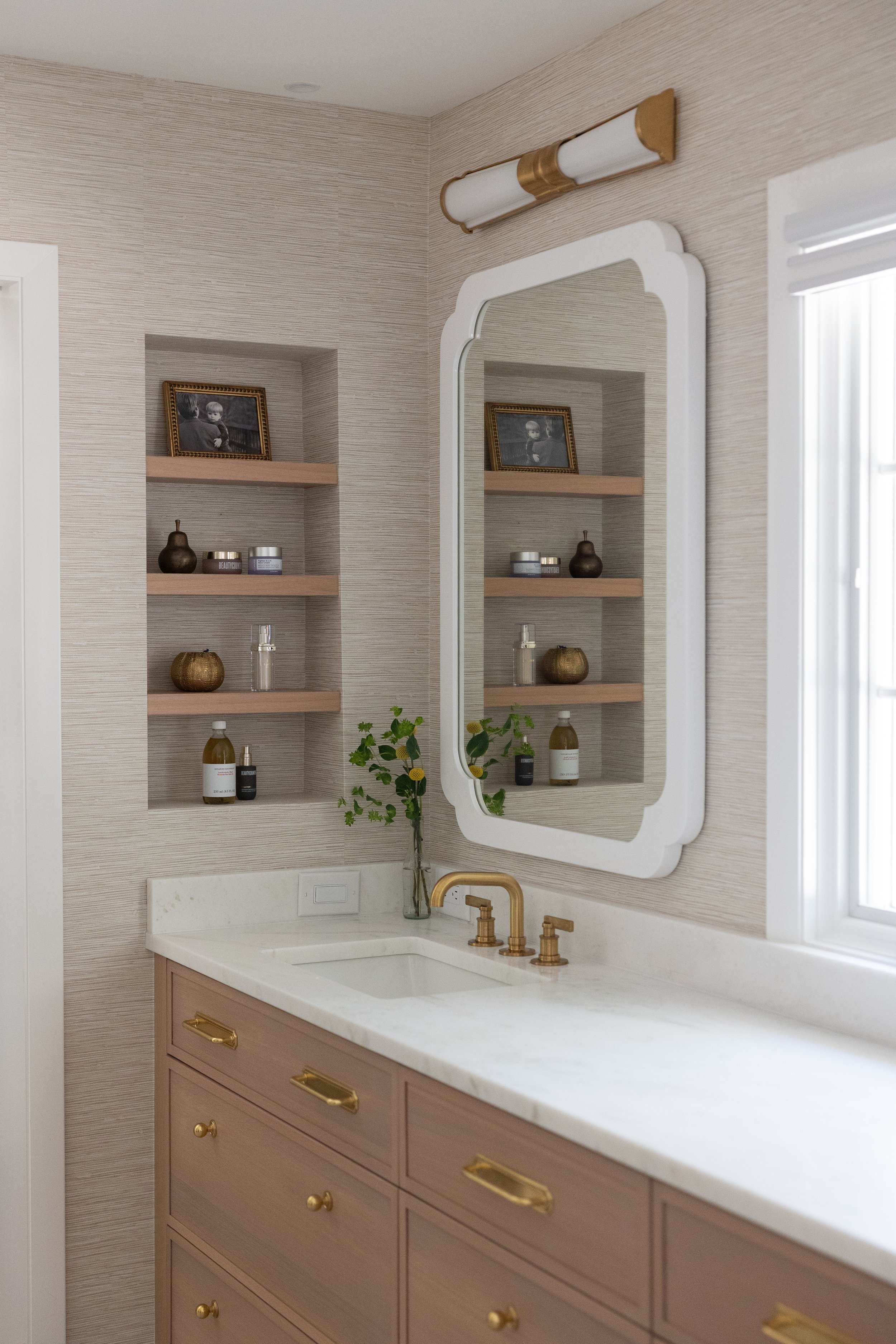 A bathroom vanity with a white countertop, gold fixtures, and a large mirror. Behind the mirror are built-in shelves with decorative items, and a small window is on the right side.