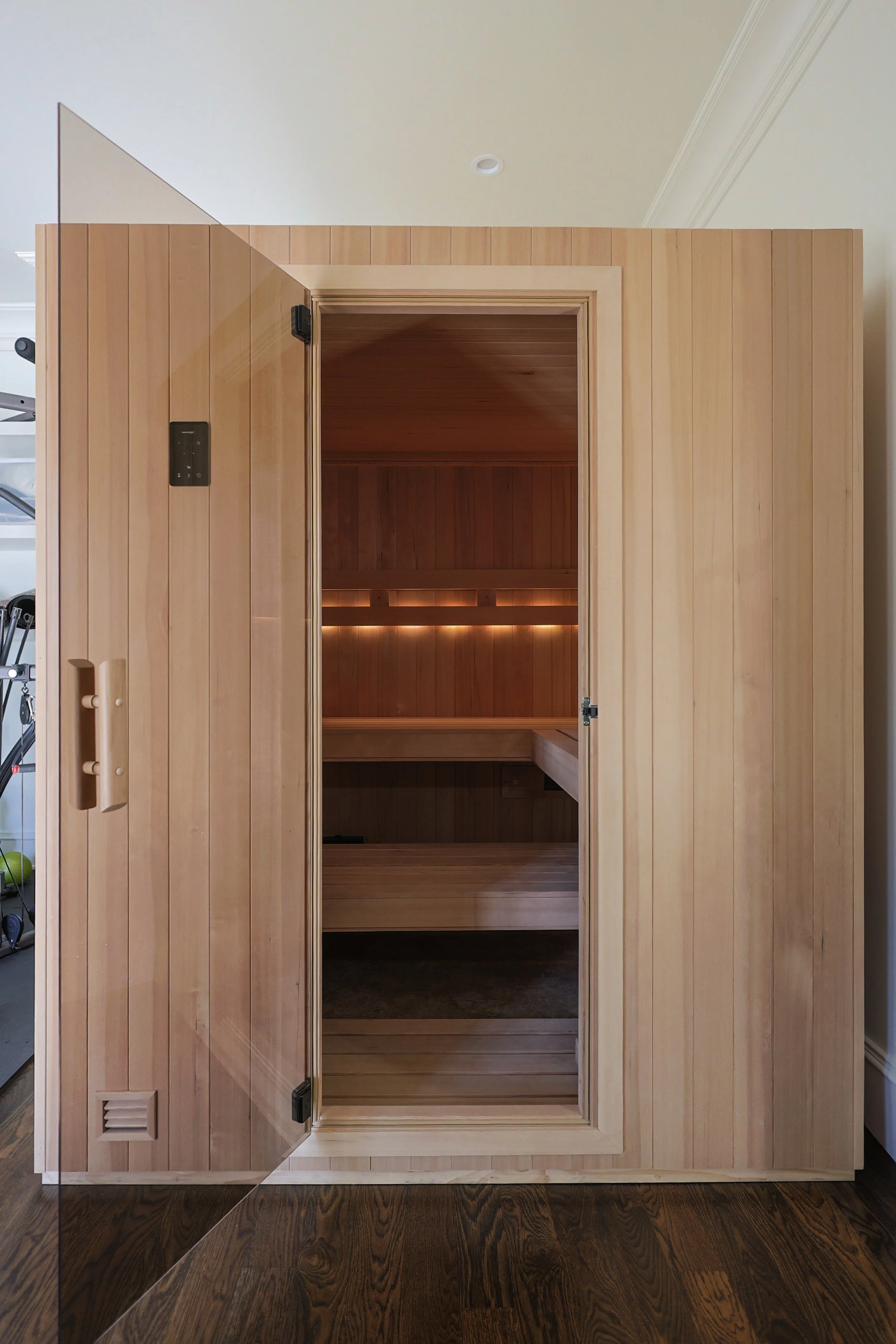 A wooden infrared sauna with an open glass door showing the interior with built-in seating and ambient lighting.