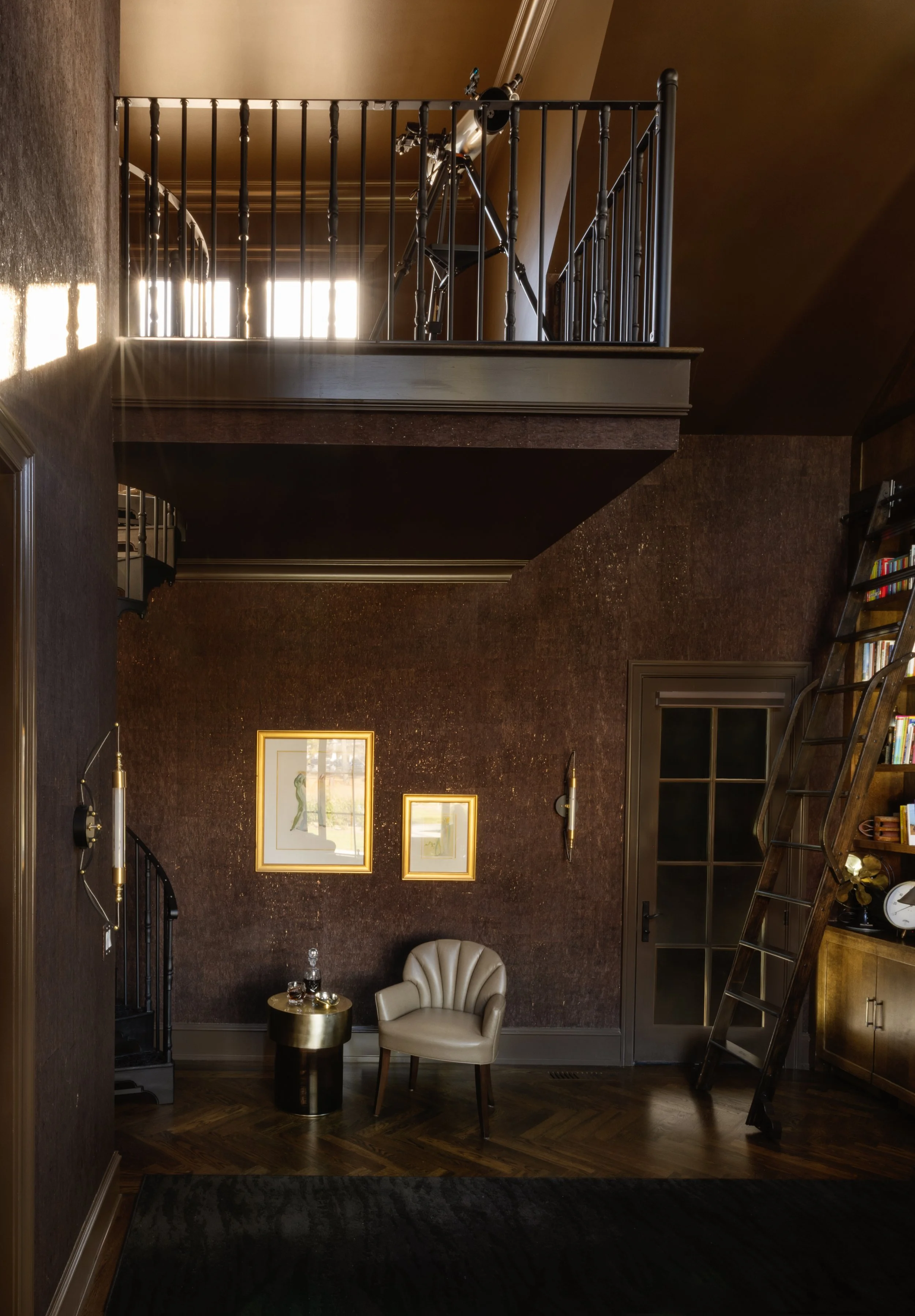 Interior of a room with dark textured walls, a beige chair, gold-framed artwork, and a birdbath-style side table with glassware. A wooden ladder leaning against a bookshelf and a glass door are visible. Balcony with telescope is seen above.
