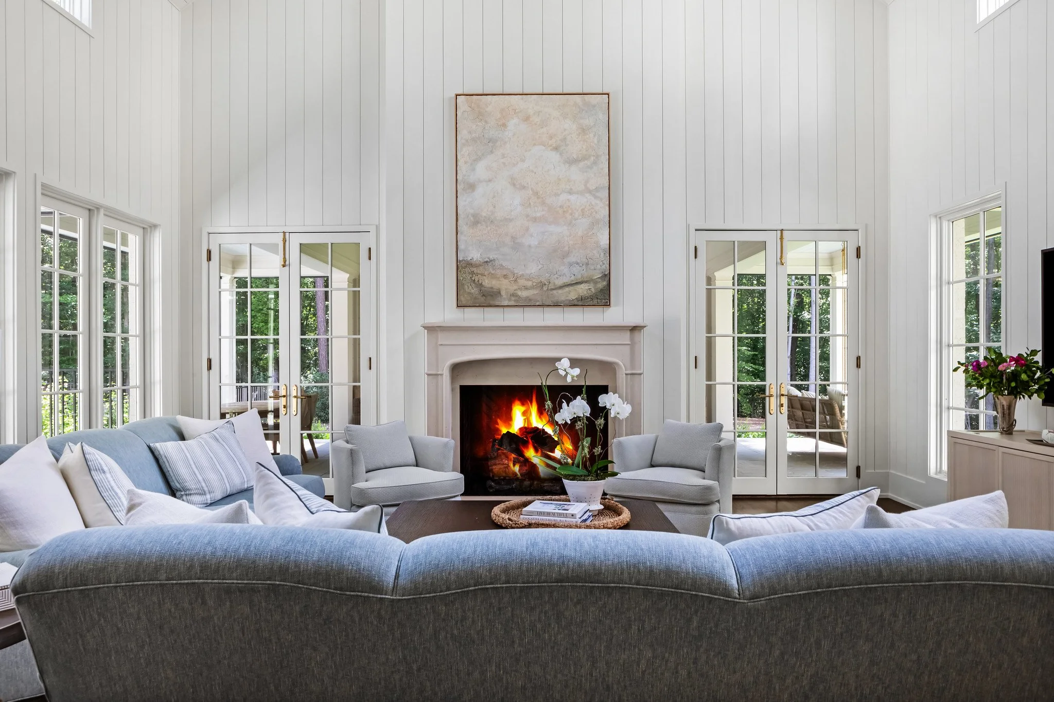 Living room with white paneled walls, a fireplace with a fire burning, surrounded by gray and white furniture, large windows and French doors, a round table with a plant, and a large abstract painting above the fireplace.