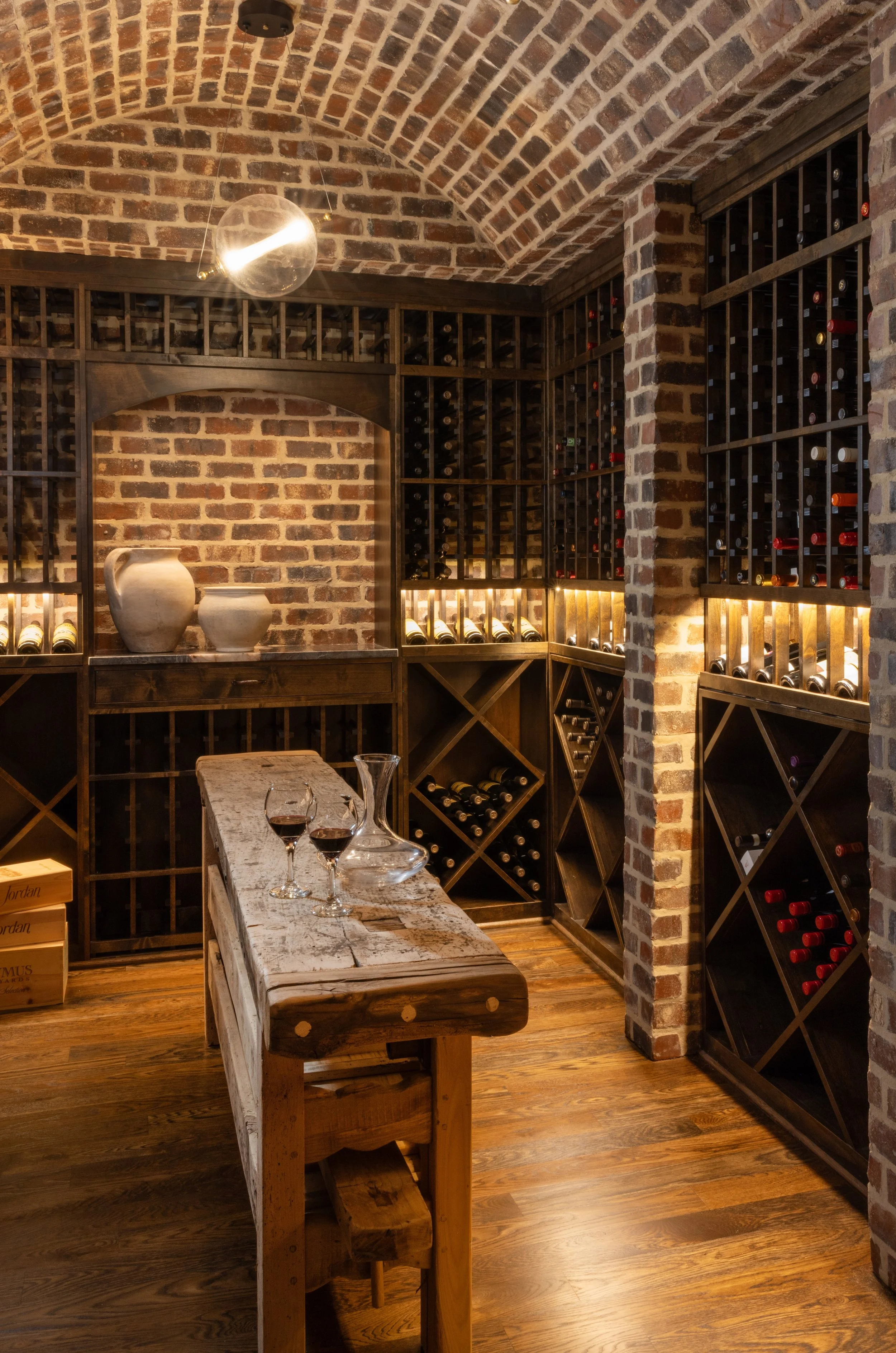 A wine cellar with brick walls and arched brick ceiling, wooden wine racks filled with bottles, a rustic wooden table with a glass decanter and two glasses of red wine, and decorative pottery on a shelf.