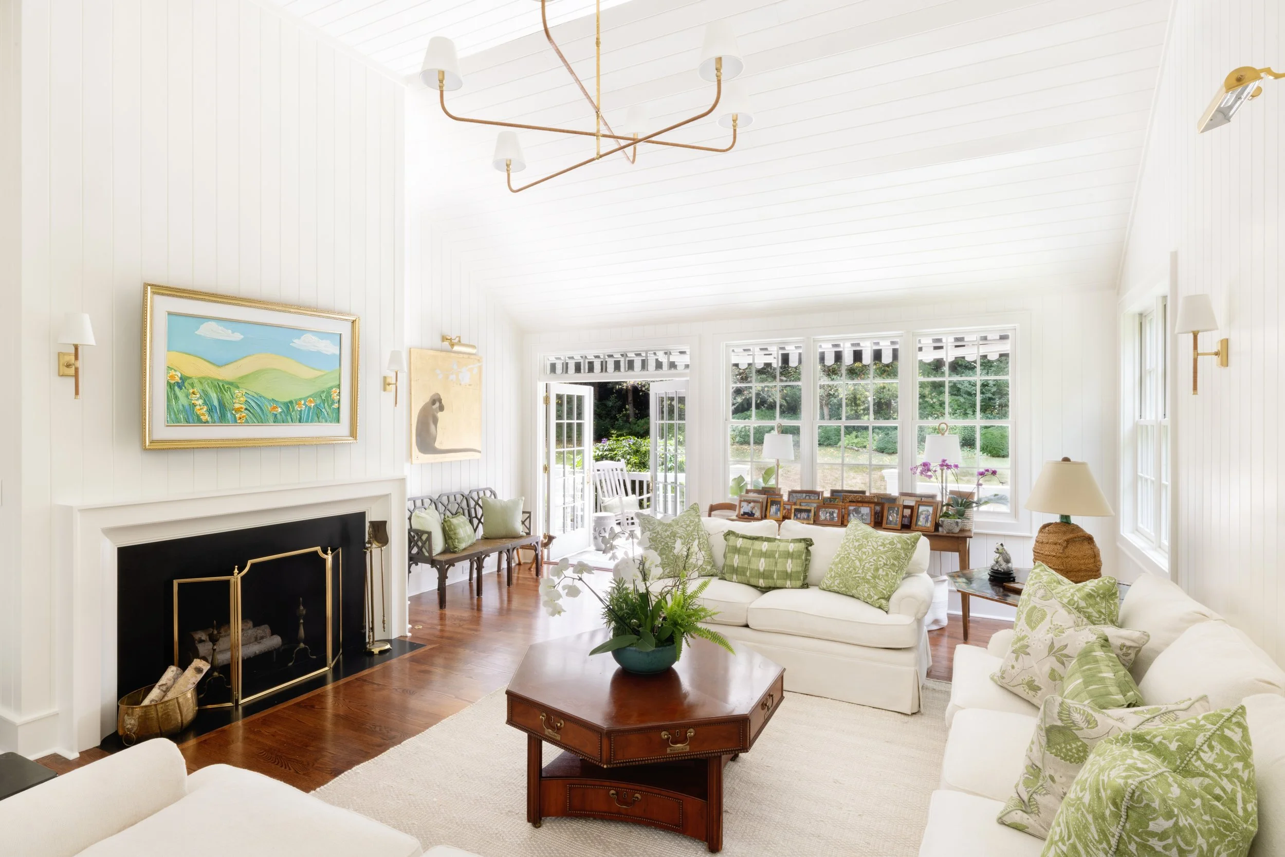 Bright living room with white walls and ceiling, hardwood flooring, and large windows. Contains white sofas with green patterned pillows, a wooden coffee table with a plant, a fireplace with logs, and framed photos on a side table. Artwork and lamps 