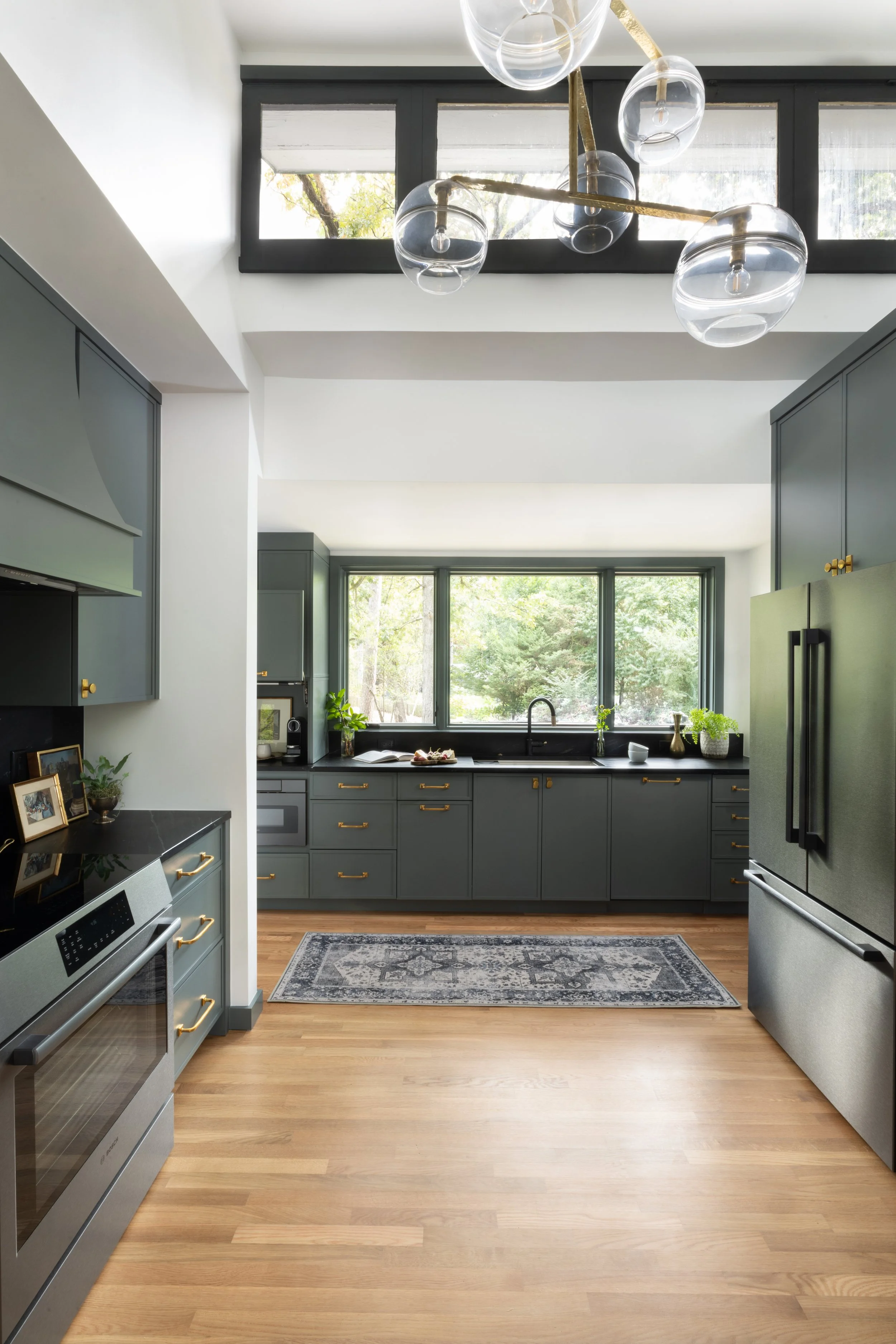 Modern kitchen with gray cabinets, black countertops, and stainless-steel appliances, including a refrigerator and oven. Large window above the sink overlooks green trees, and a contemporary chandelier hangs from the ceiling. The floor is hardwood, a
