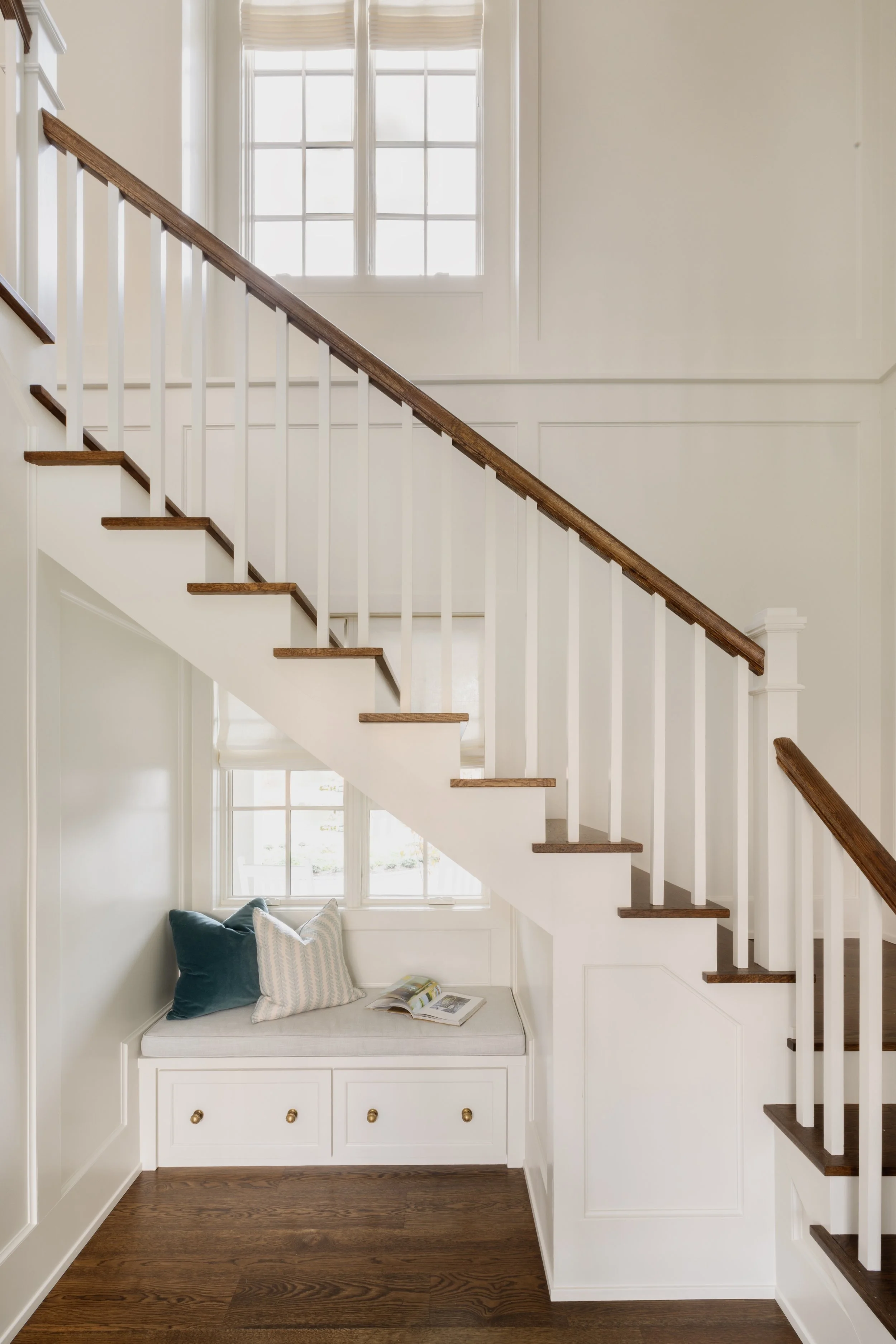 A staircase with white risers, dark wooden treads, and matching dark wooden handrail. A built-in white bench with drawers, decorated with pillows and an open magazine, is situated at the base of the staircase beneath a large window. The walls are pai